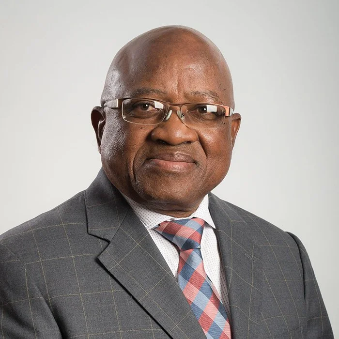 A professional African American man wearing glasses, a plaid tie, and a dark checked suit against a neutral background.