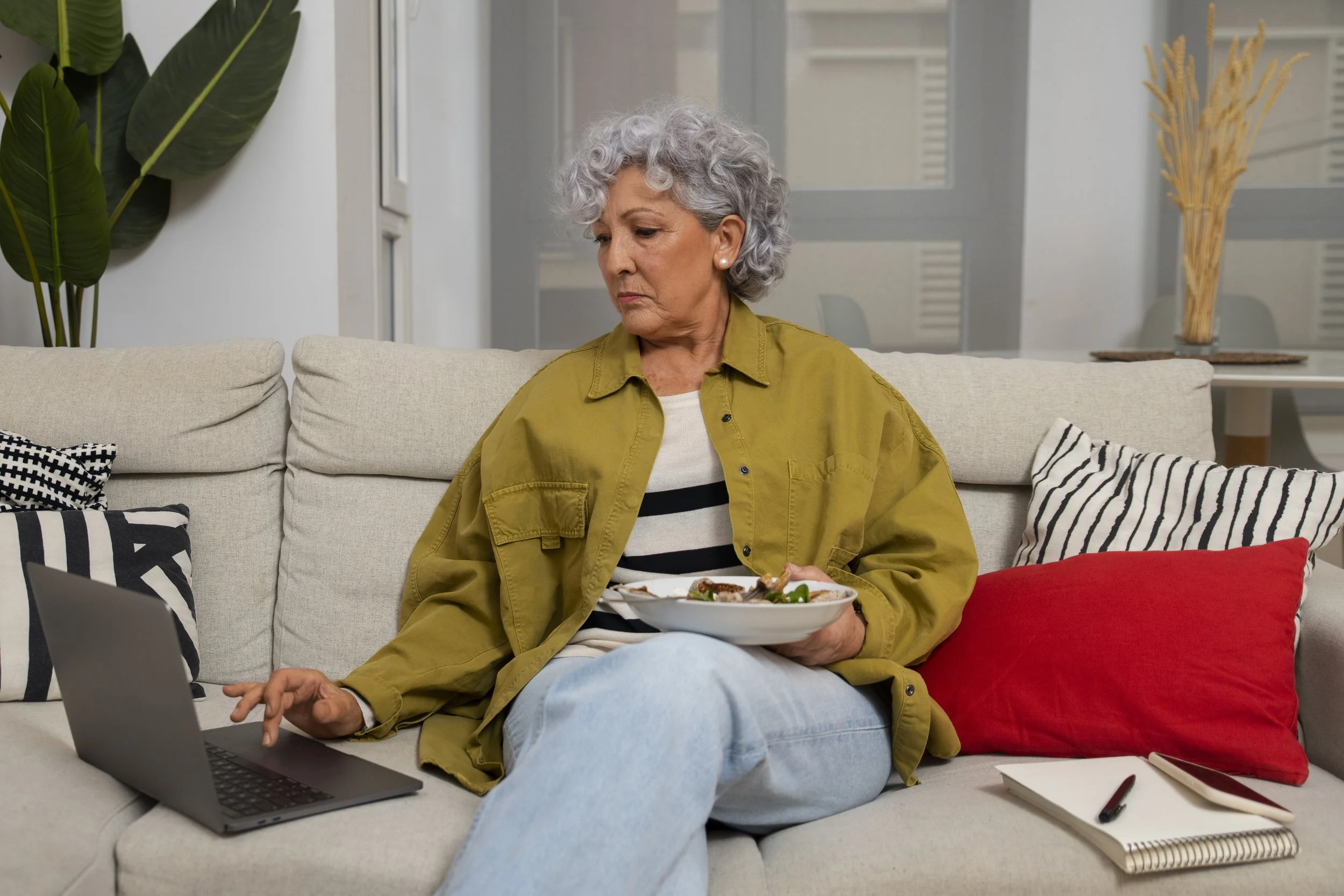 An elderly woman with gray curly hair, wearing a yellow jacket and striped shirt, sitting on a beige sofa in a living room. She is holding a plate of food in one hand and using a laptop with her other hand. There are patterned pillows, a white notebook, and a pen on the sofa, with green plants and a vase in the background.