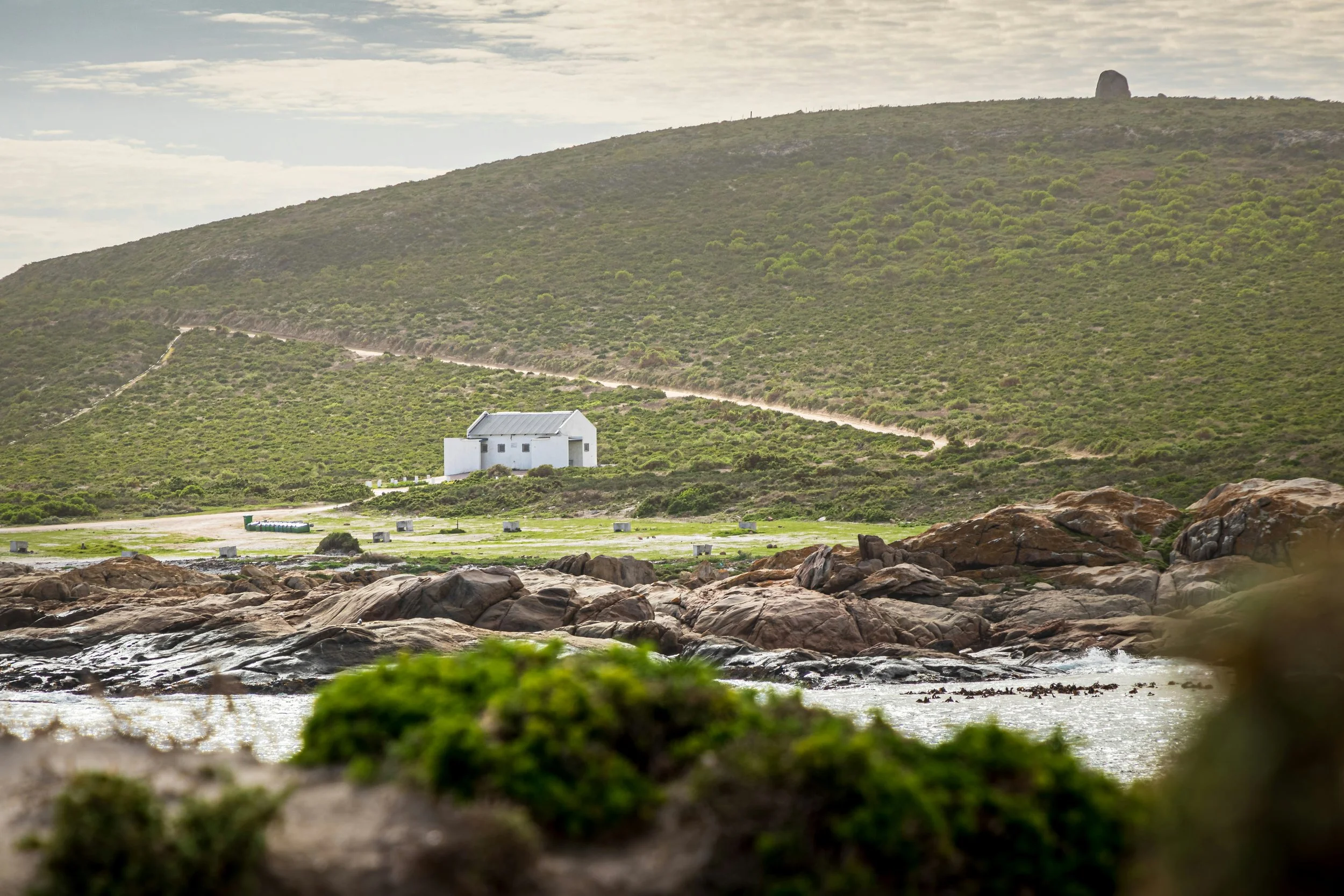 A small white house on a grassy area near rocky shoreline, with a hillside and path in the background under a cloudy sky.