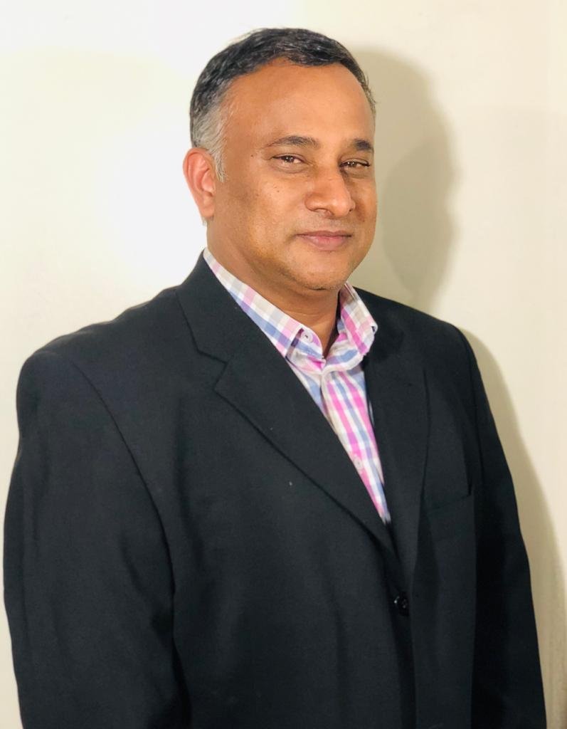 A man with short dark hair and a slight smile, wearing a black blazer over a pink, purple, and white checkered shirt, standing against a plain white wall.