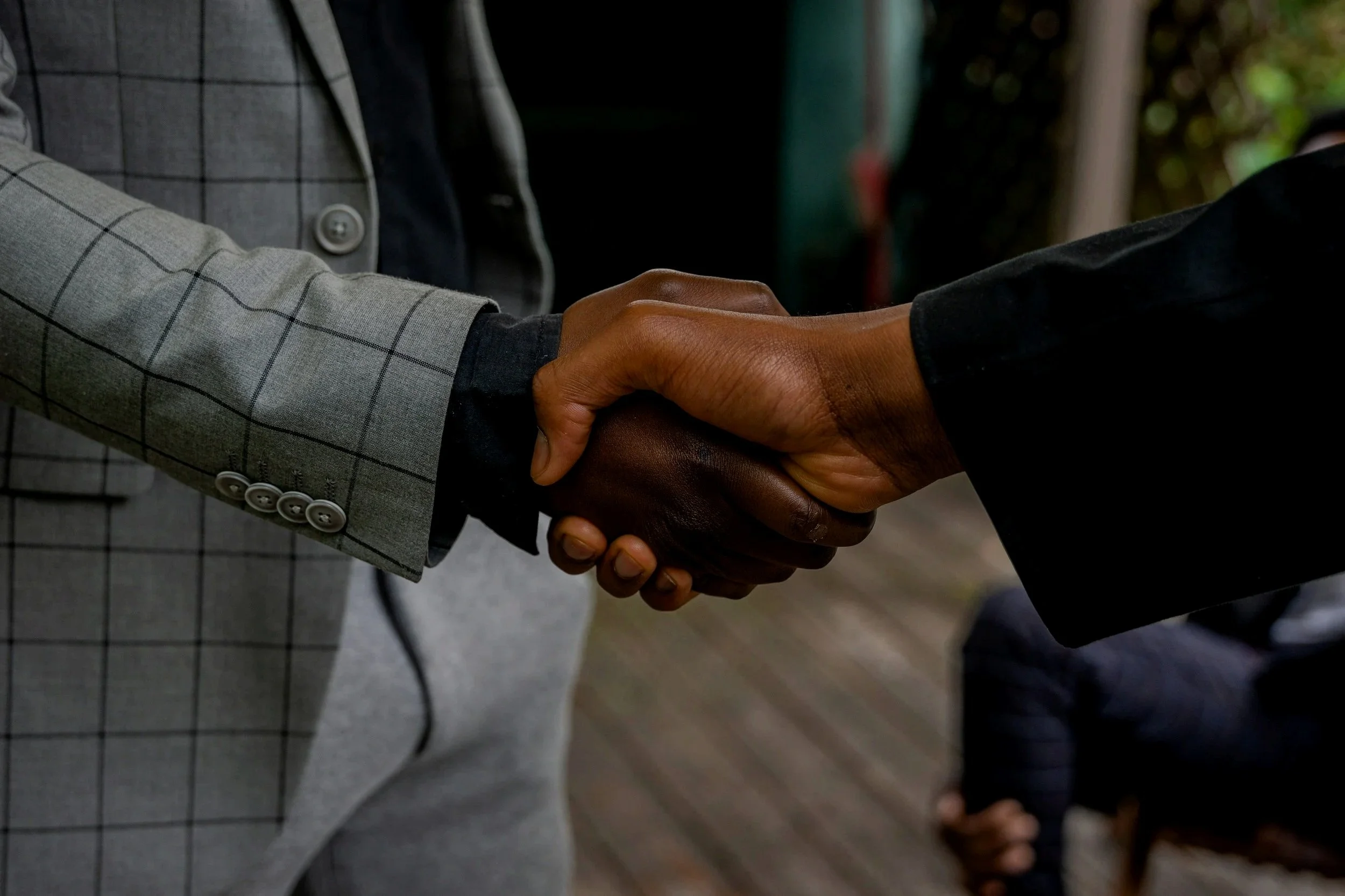 Two people shaking hands, one wearing a grey checkered suit and the other wearing a black suit.