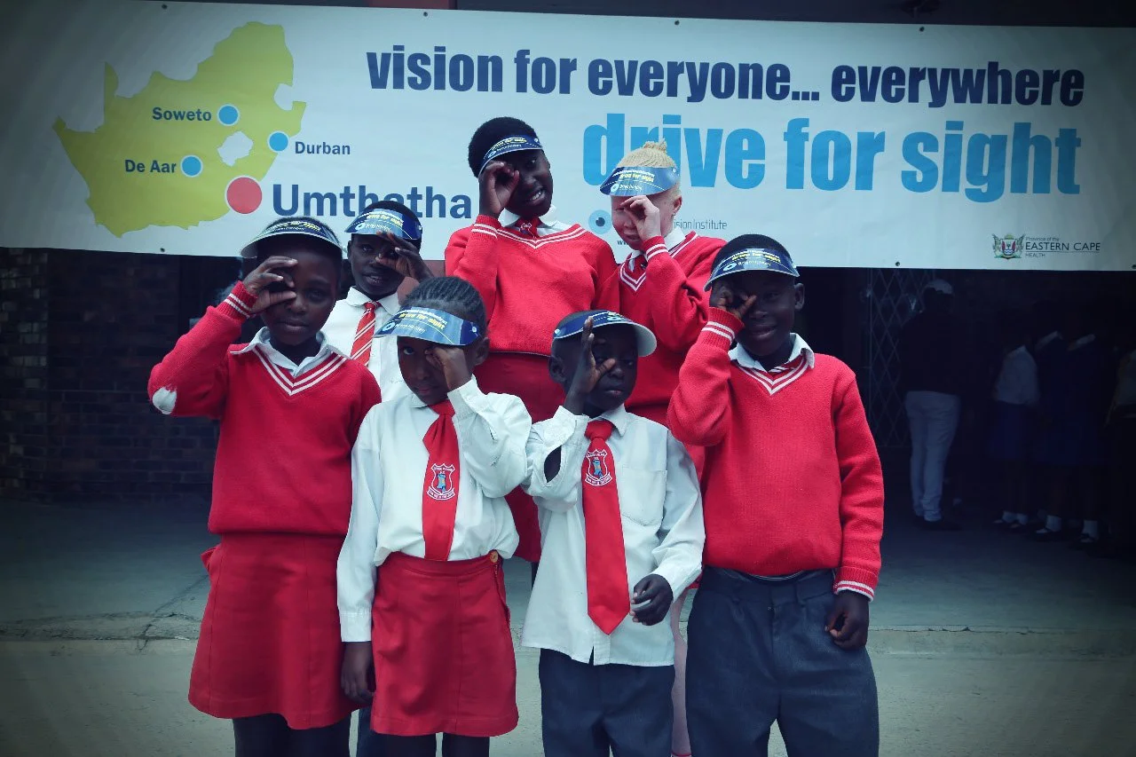 Six school children in uniforms participating in a sight-driving awareness event, saluting and wearing branded helmets, standing in front of a banner with a map of South Africa and the words 'vision for everyone... everywhere, drive for sight'.