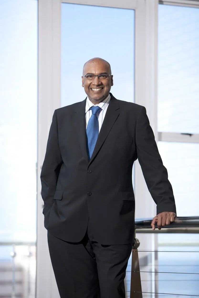 A smiling man in a black suit, white shirt, and blue tie standing indoors by a railing in front of large windows.