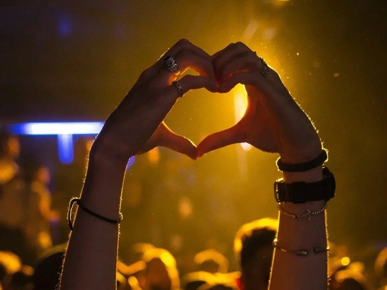 Hands forming a heart shape against a warm, glowing background at a crowded event.