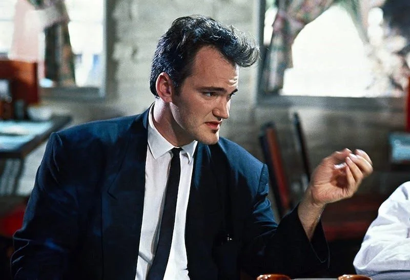 Quentin Tarantino man in a navy blue suit and tie sitting at a table in a restaurant, looking down with a serious expression, while gesturing with his right hand.