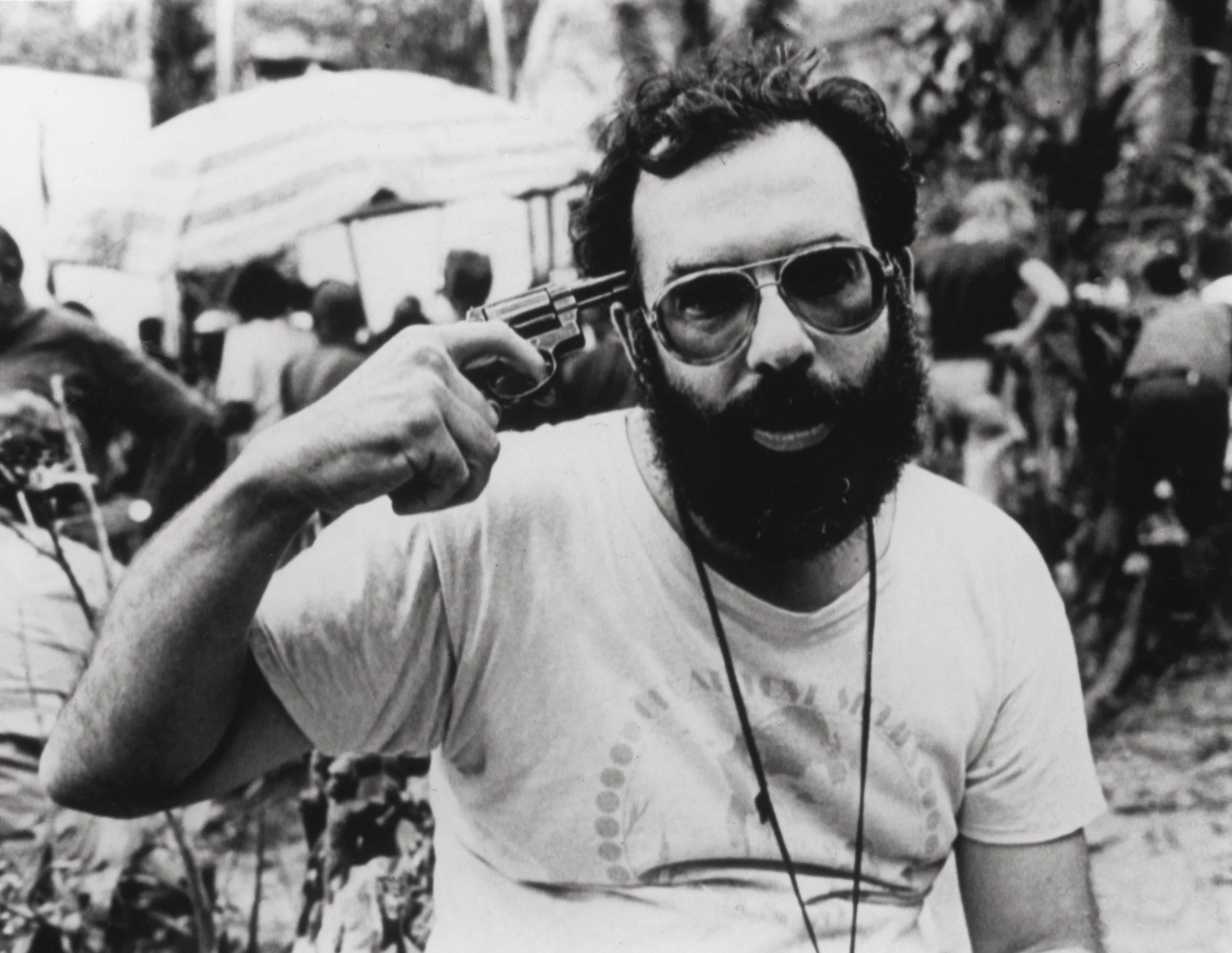 Francis Ford Coppola man with a beard and sunglasses holding a handgun to his head, at an outdoor gathering with tents and people in the background.