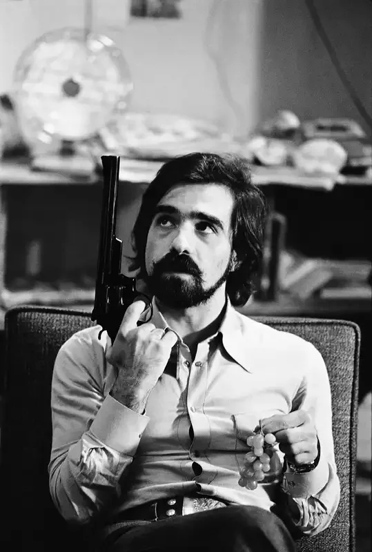 Martin Scorsese man with dark hair and a beard, wearing a light-colored shirt, sitting on a chair, holding a handgun to his temple with his right hand, and holding grapes in his left hand, in a room with shelves and objects in the background.