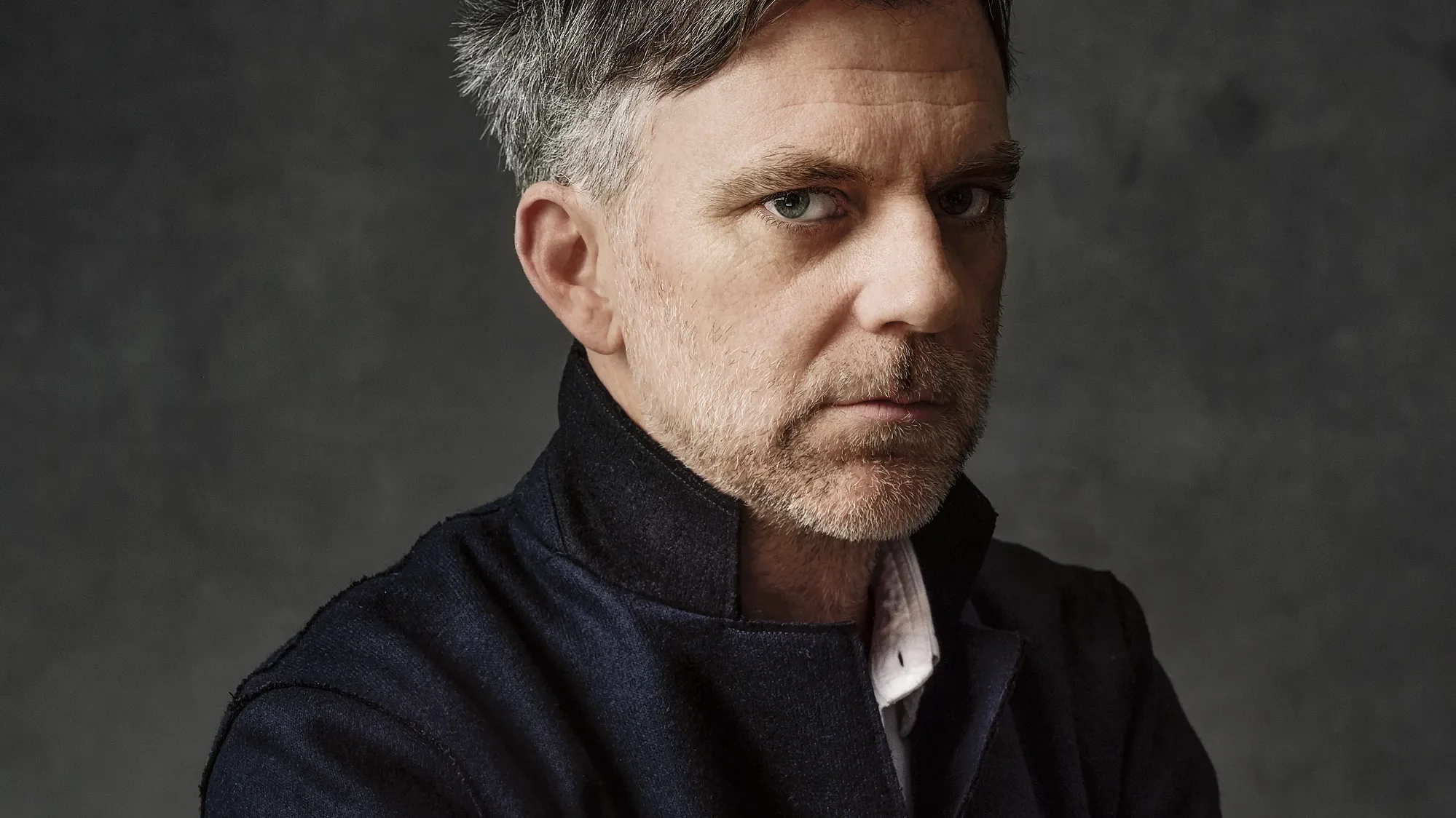 A close-up portrait of Paul Thomas Anderson middle-aged man with short gray hair and a beard, wearing a dark jacket, looking slightly to the side with a serious expression against a dark background.