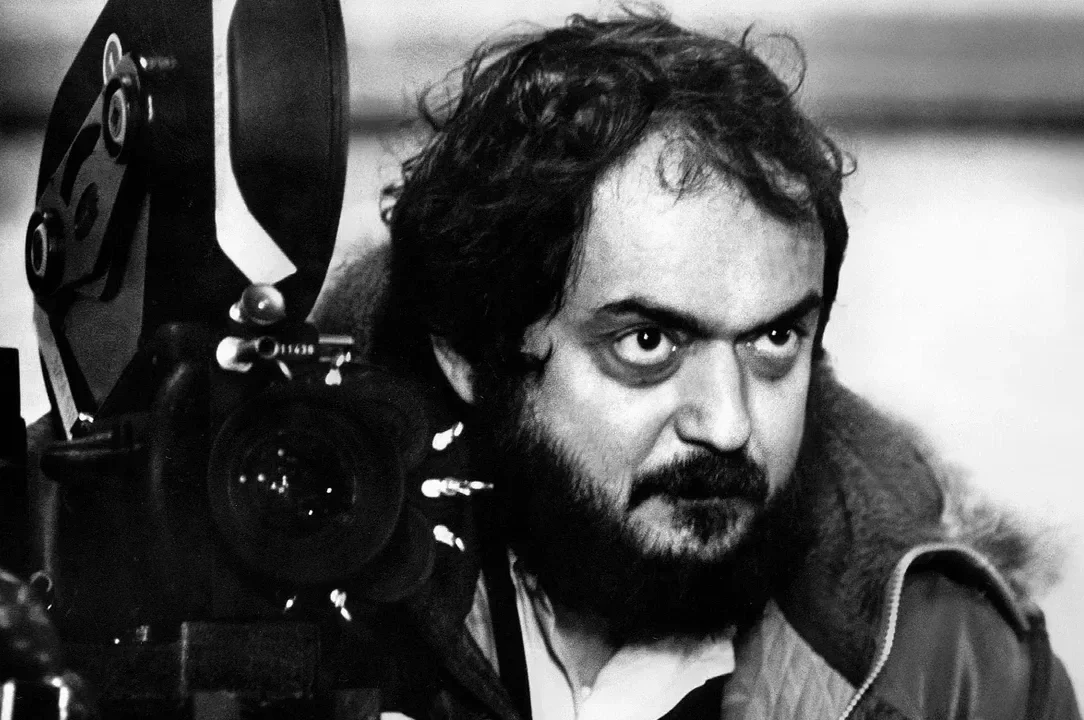 Black and white photo of Stanley Kubrick man with curly hair and beard, behind film camera equipment, looking intently to the side.