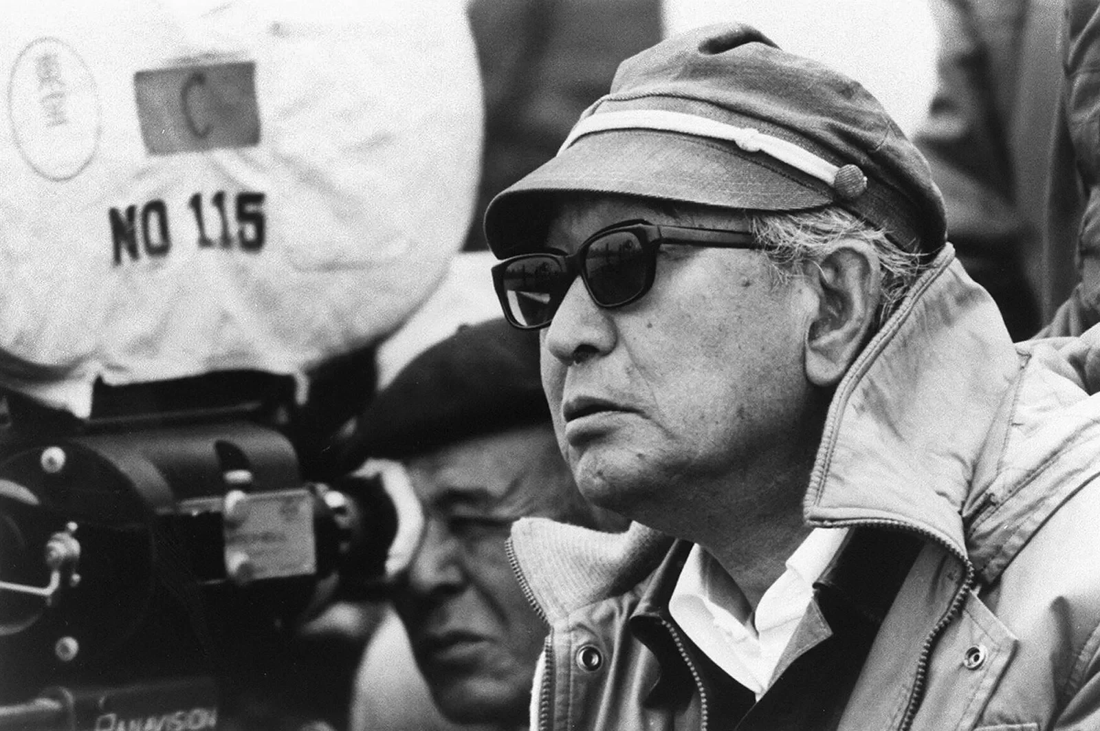 Akira Kurosawa man wearing sunglasses, a cap, and a jacket is standing next to a person operating a film camera.