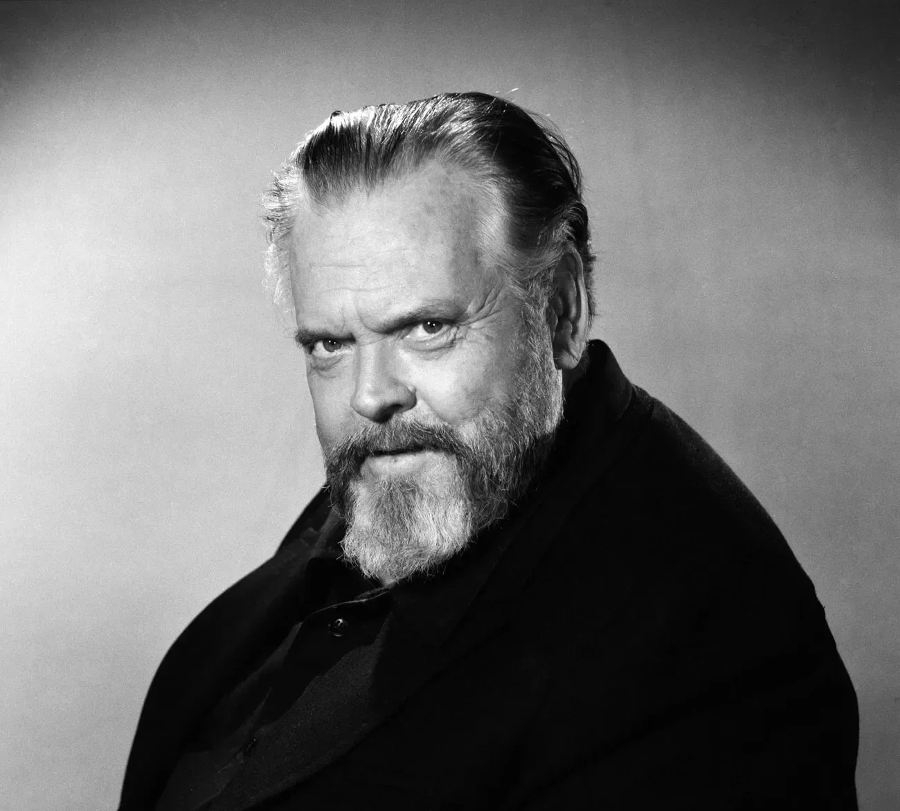 Black and white portrait of Orson Welles bearded man with styled hair, looking slightly to the right of the camera with an intense expression.