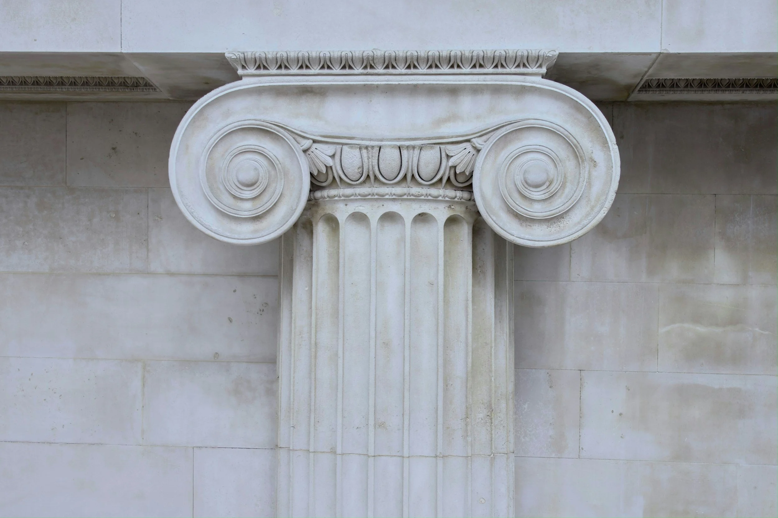 Close-up of a white classical Ionic column capital with volutes and floral details, part of a building facade.