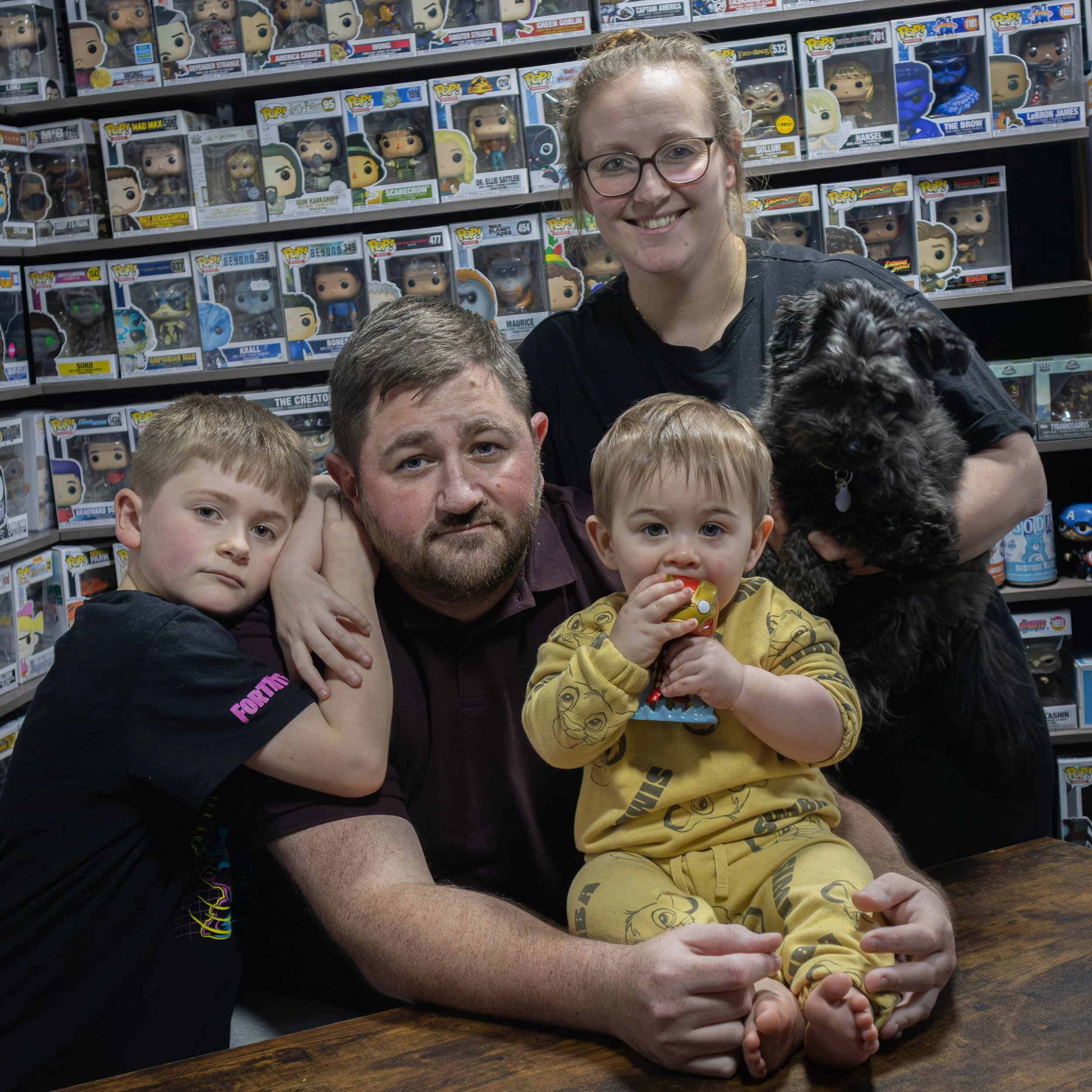 A family of five with a dog in a toy store surrounded by shelves of Funko Pop! collectibles. The family includes two young boys, an adult man, an adult woman, and a toddler girl sitting on the man's lap. The boy on the left wears a black T-shirt, the toddler girl in yellow pajamas is holding a toy, and the woman is holding a black dog. Everyone is smiling or looking at the camera.