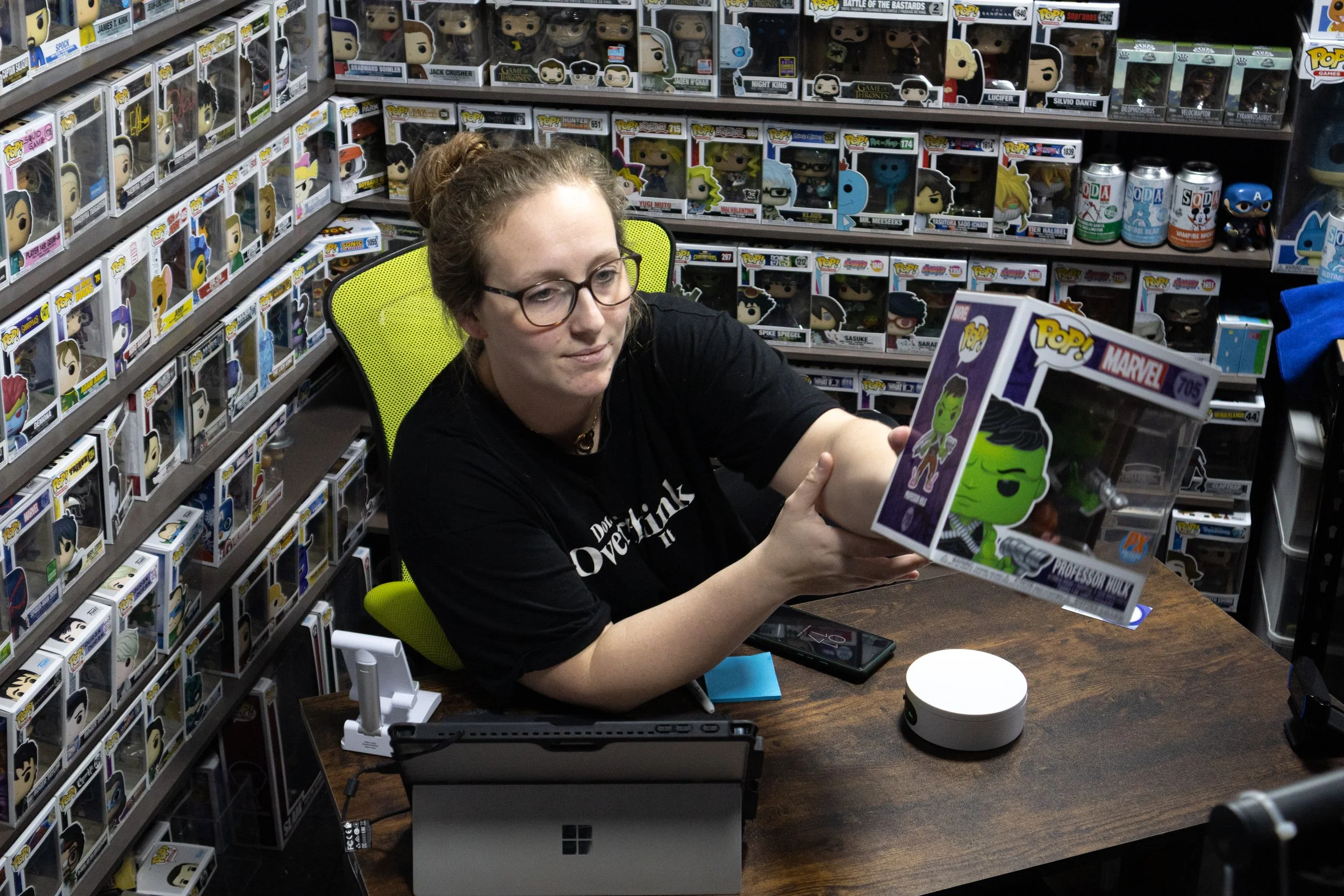 A woman with glasses and a bun hairstyle sitting at a desk, holding a boxed Funko Pop! figure of Professor Hulk from Marvel. The background is filled with shelves of collectible figures.