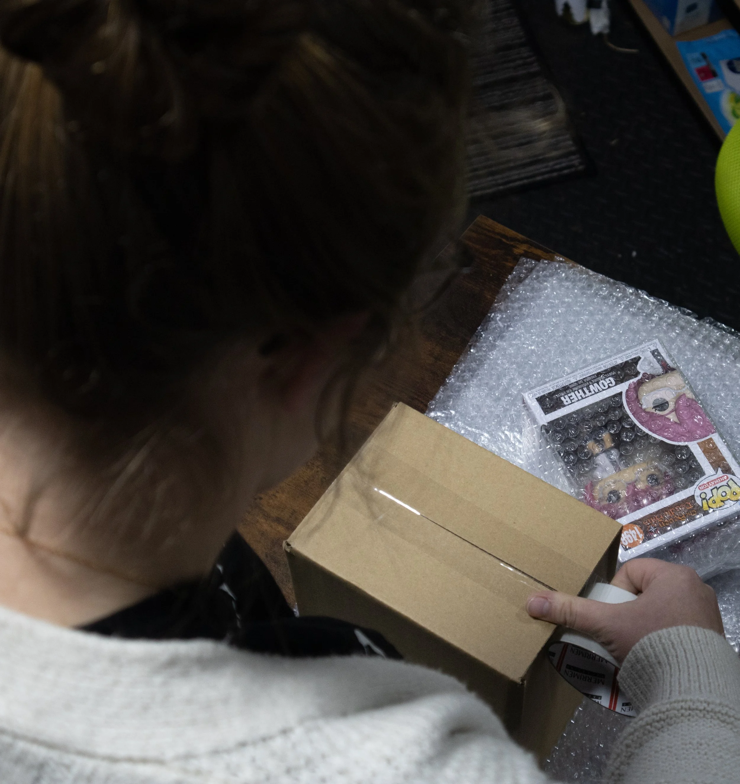 A person opening a cardboard box on a table with a Funko Pop figurine in bubble wrap, next to a comic book and packing materials.
