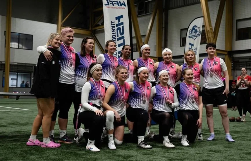 A group of women and girls in sports uniforms posing for a team photo indoors, with medals around their necks, standing on a green sports field.
