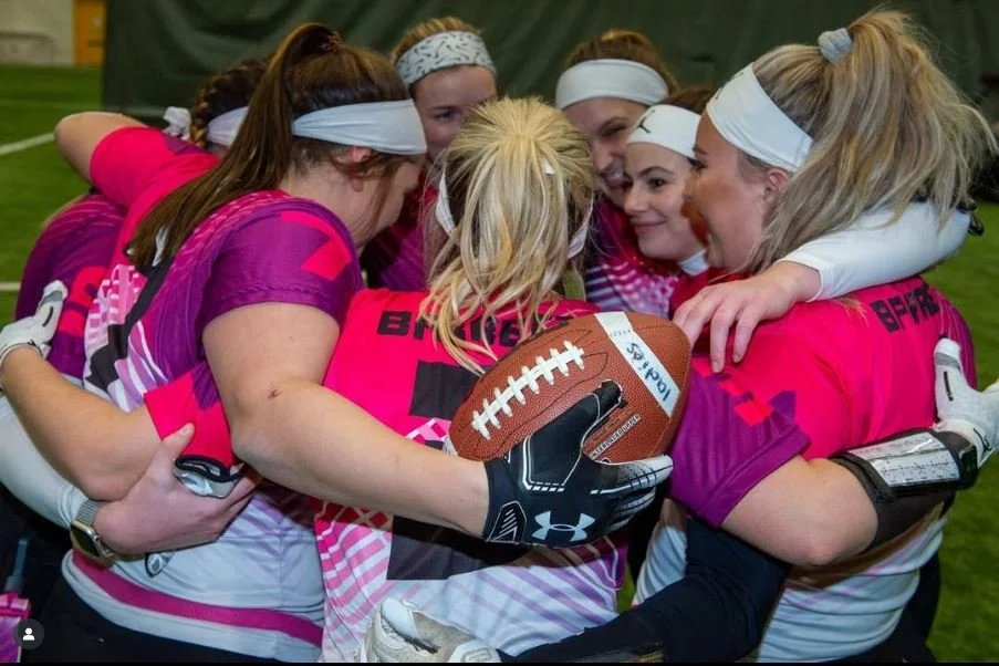 A group of young female football players wearing pink and purple jerseys, gathered in a huddle, holding a football, with smiles and enthusiasm.