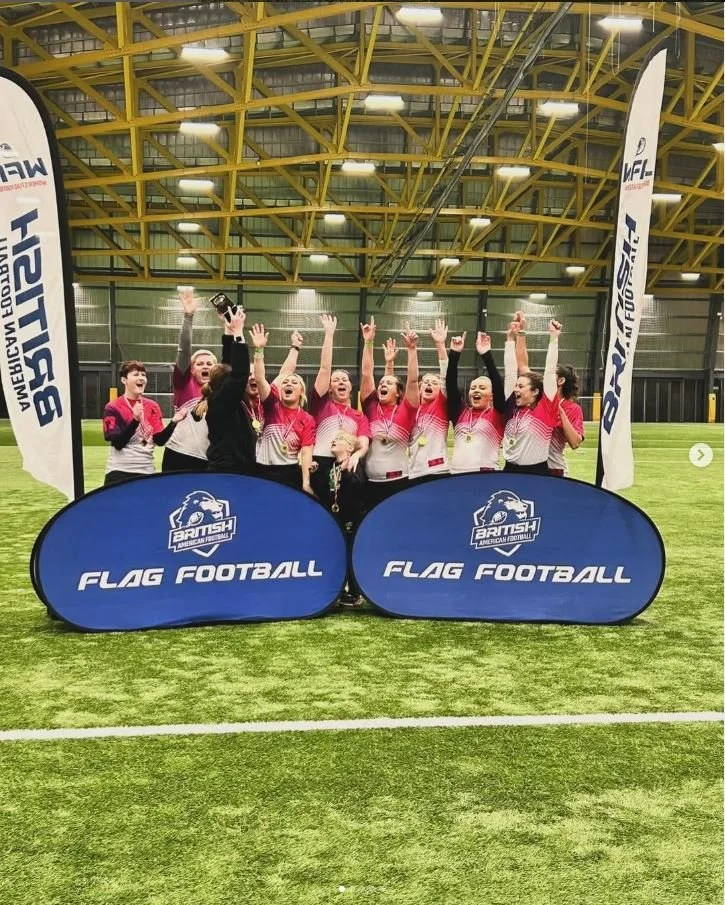 A group of female flag football players in pink and white jerseys celebrating indoors on a green turf field with banners that say 'Flag Football' and 'BRITISH American Football'.