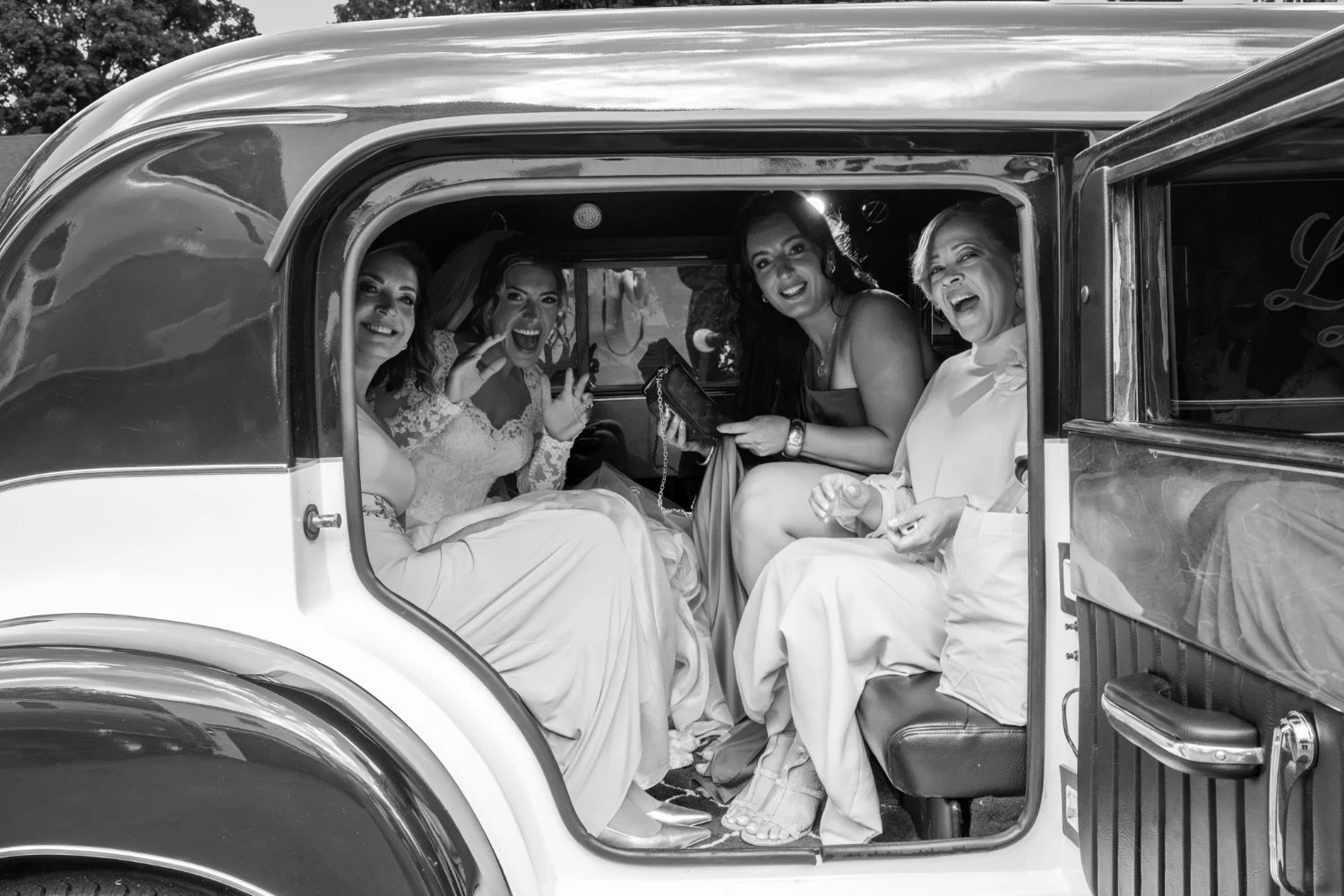 Four women sitting inside a vintage car, smiling and laughing, dressed in formal attire, possibly at a wedding.