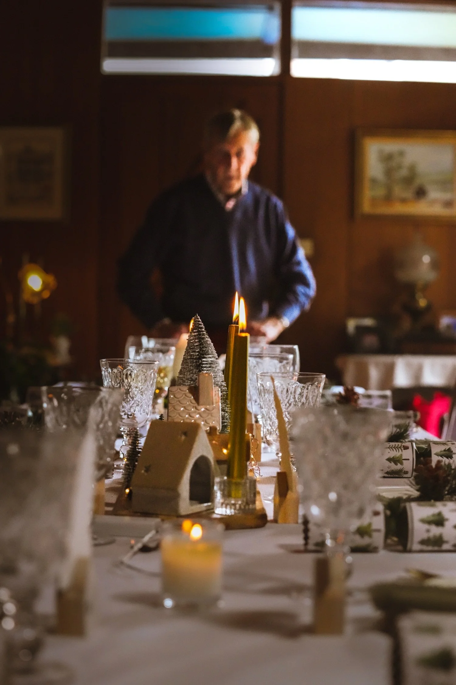 A table decorated for Christmas with candles, small houses, and ornaments. In the background, a man stands in front of a wooden wall with framed pictures, illuminated by natural light from a window.
