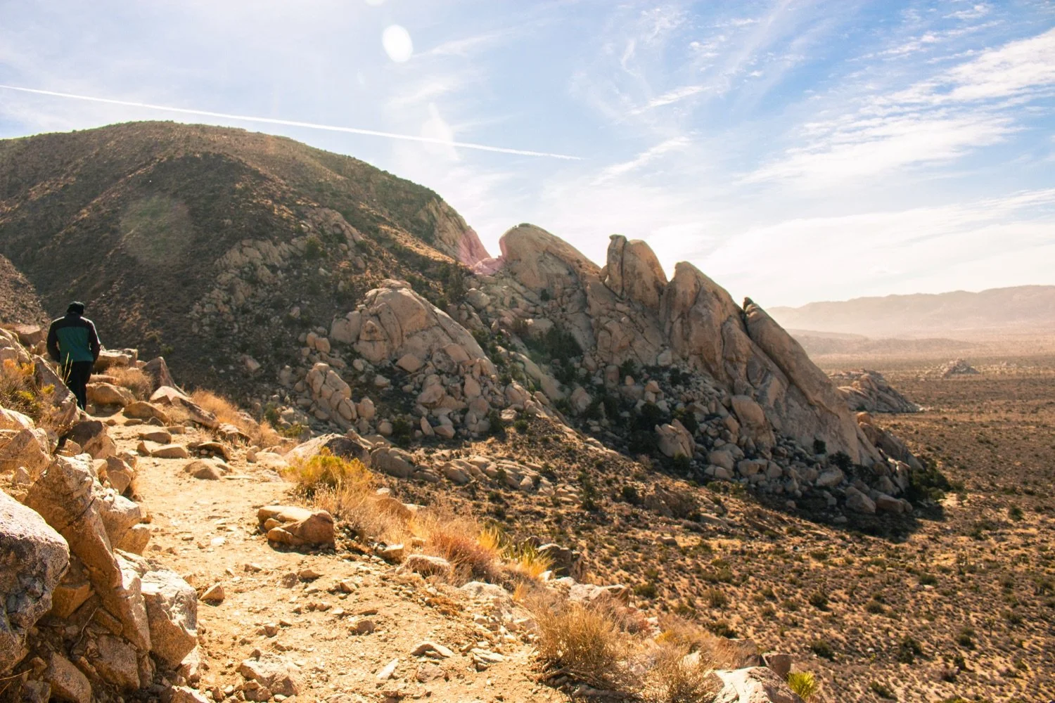A hiker walking along a rocky desert trail with large boulder formations and a mountainous landscape in the background under a partly cloudy sky.