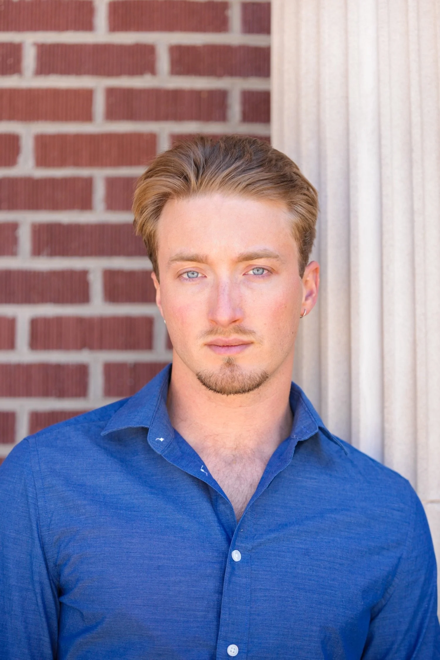 A young man with styled blond hair, blue eyes, and light skin, wearing a blue button-up shirt, stands in front of a background with a red brick wall and white vertical siding.