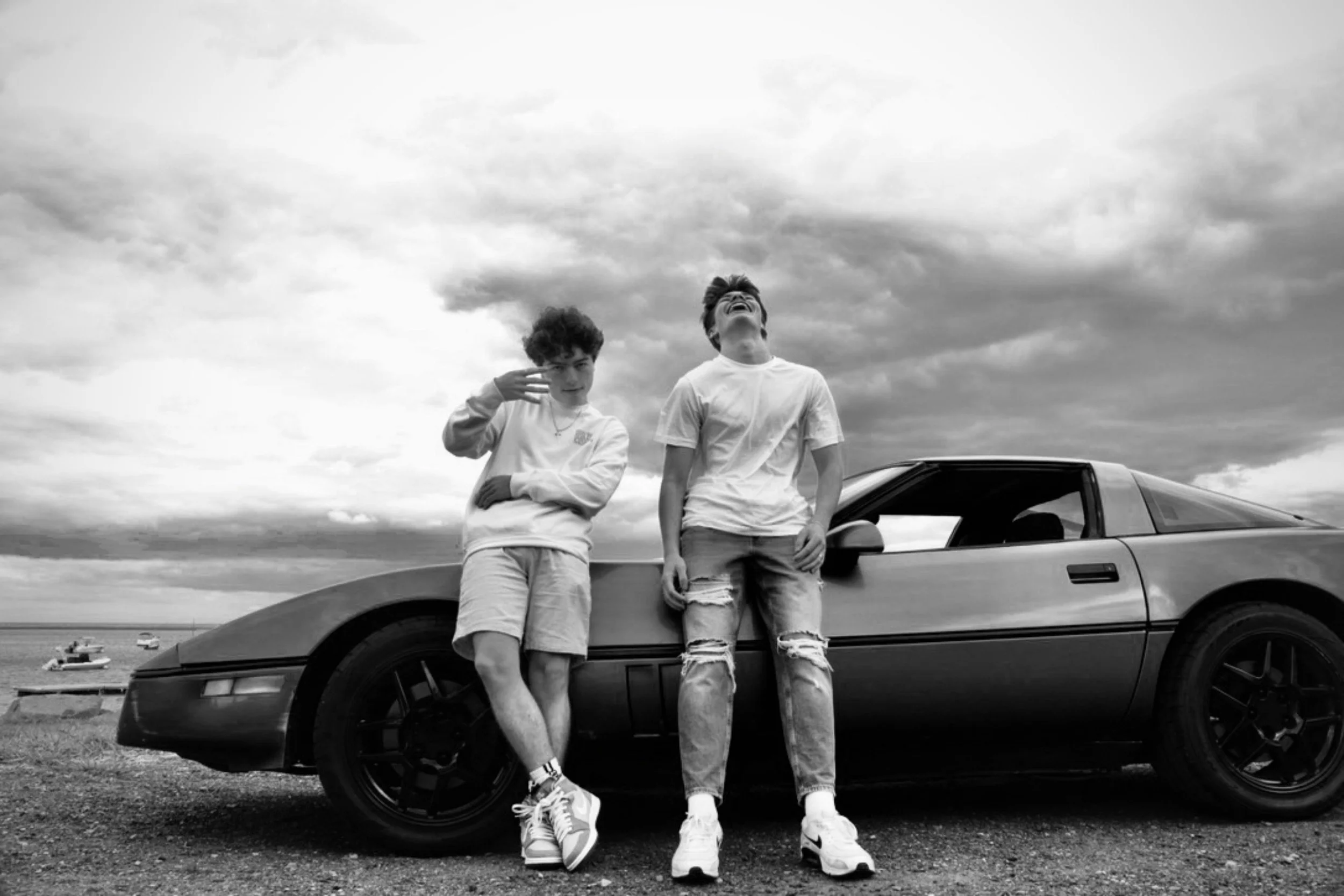 Two young men leaning against a car with a cloudy sky in the background. One is making a peace sign, the other is laughing. The scene appears to be at a beach or open area with water and boats visible in the distance.