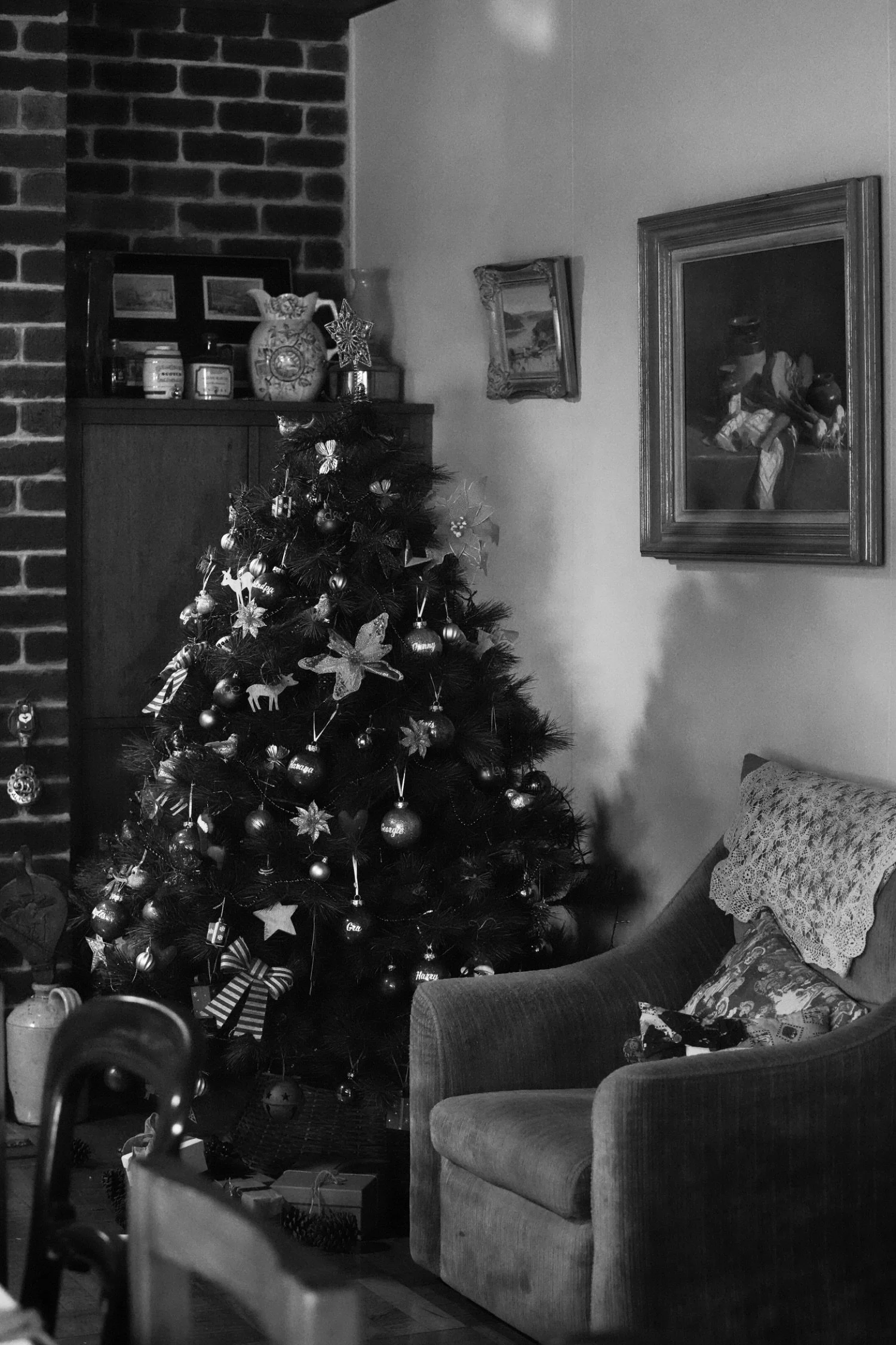 A decorated Christmas tree with ornaments, ribbons, and lights in a living room, with a wooden cabinet with decorative items, artwork on the wall, a sofa with a lace pillow, and a brick fireplace in the background.