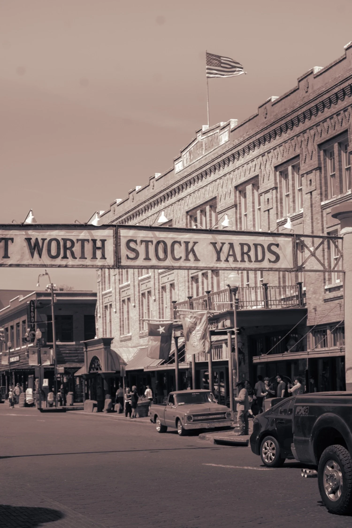 Downtown street scene with vintage cars parked and people walking, buildings with awnings, and the American flag flying atop a brick building.