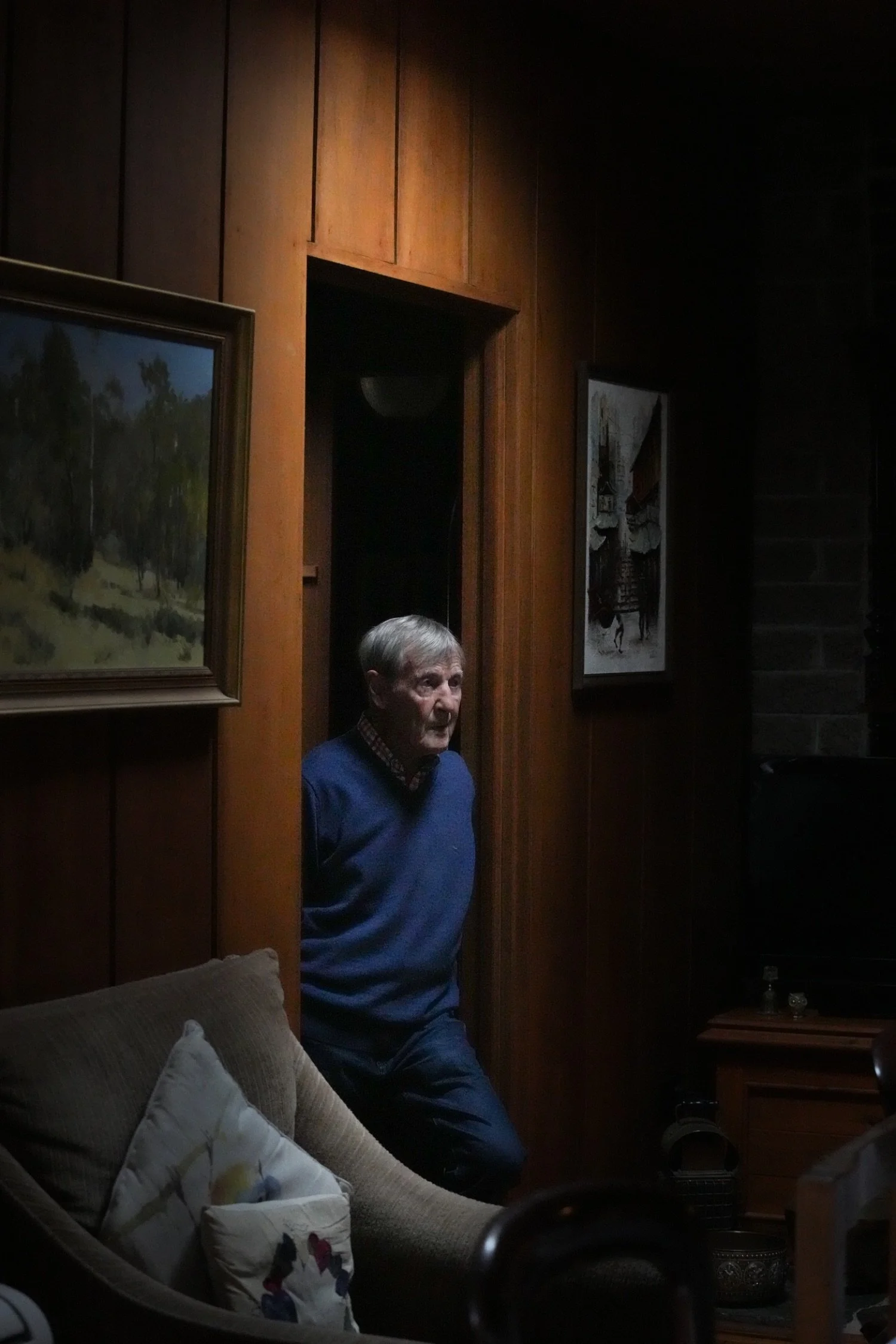 An elderly man with gray hair wearing a blue sweater, standing in a dark room with wood-paneled walls, framed paintings, and a beige sofa with cushions.