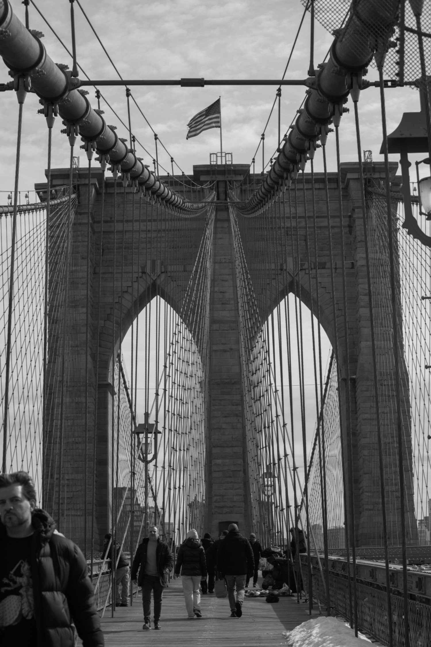 View of the Brooklyn Bridge with pedestrians walking on the bridge, and an American flag on top of the bridge structure. The photo is in black and white.