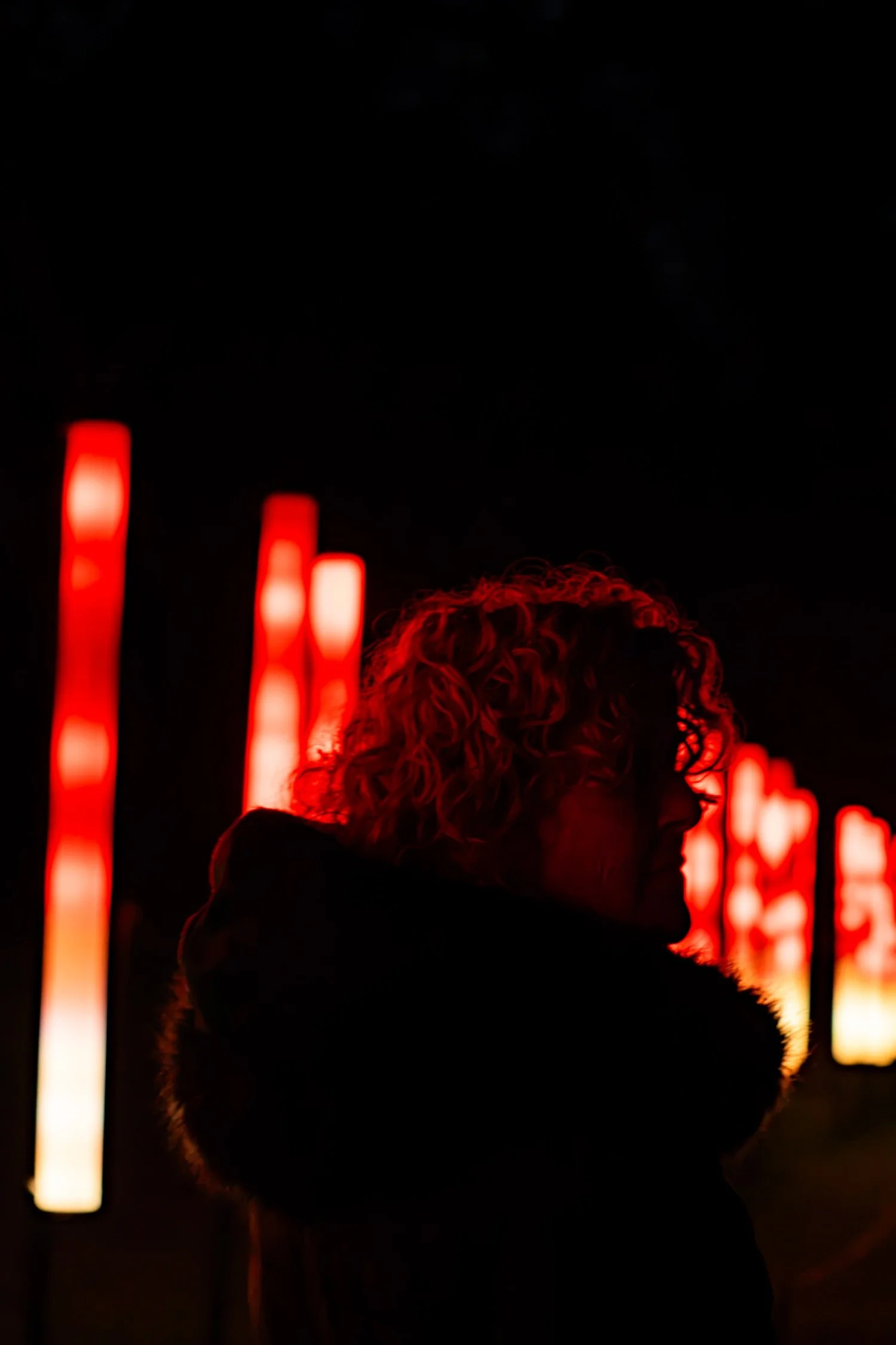 Person with curly hair standing in front of illuminated red signs at night, with silhouettes visible.