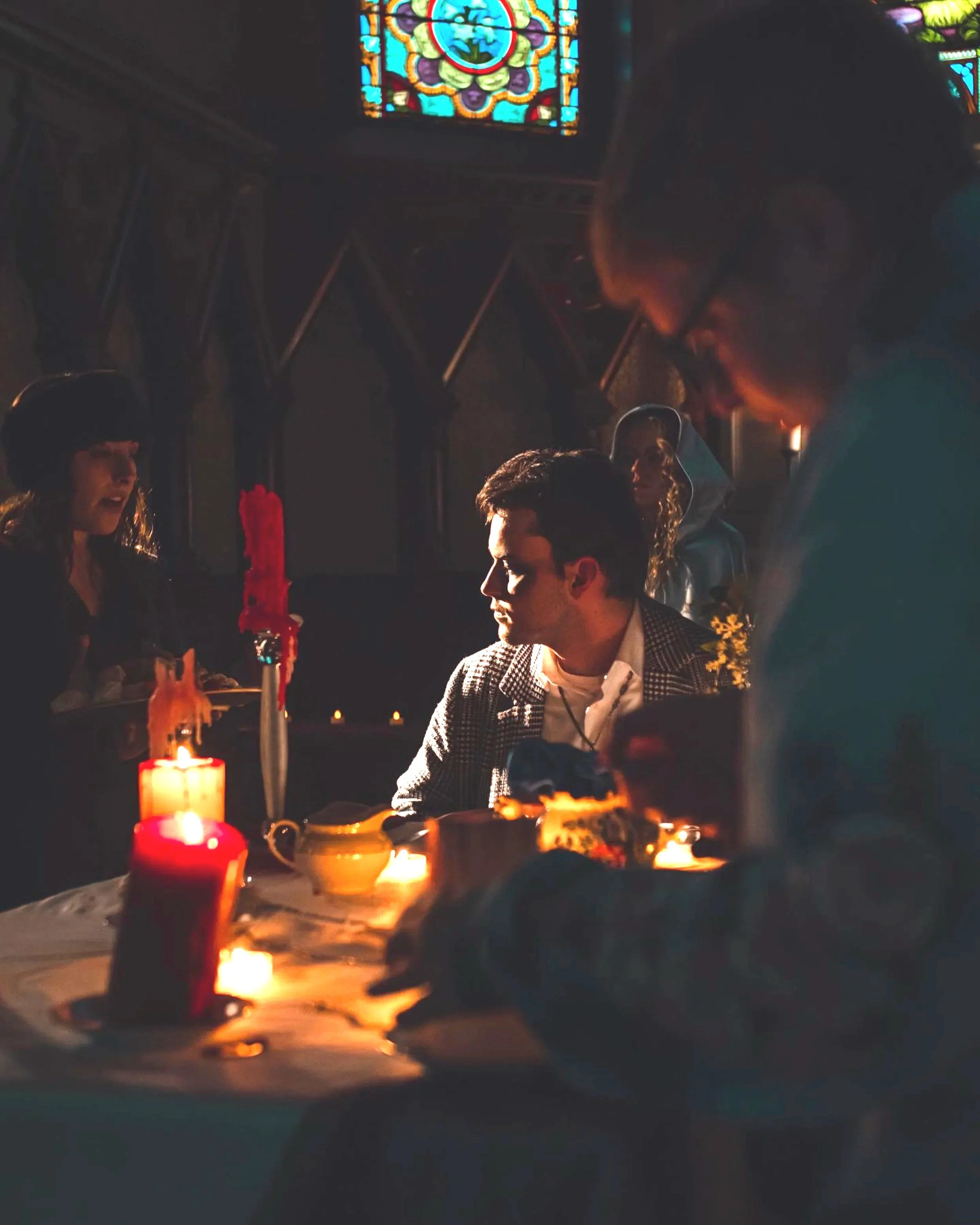 People gathered indoors around a table lit by candles, with stained glass windows visible in the background.