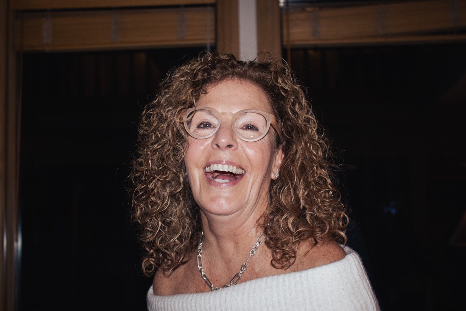 A woman with curly hair wearing glasses, a white off-the-shoulder top, and a chain necklace, smiling and laughing.