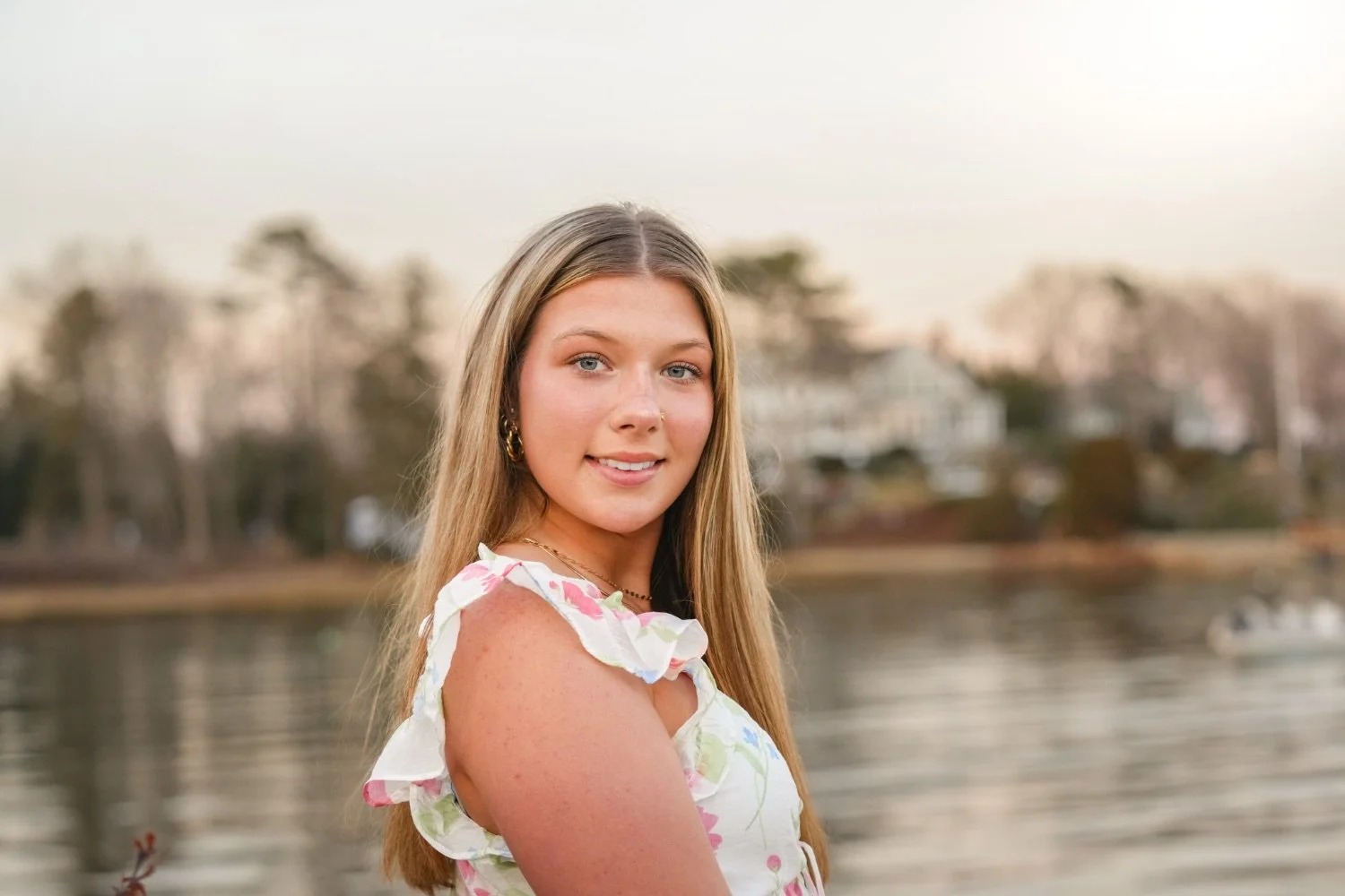 A young woman with long blonde hair, blue eyes, and wearing a floral dress, standing outdoors near a body of water with houses and trees in the background during sunset.