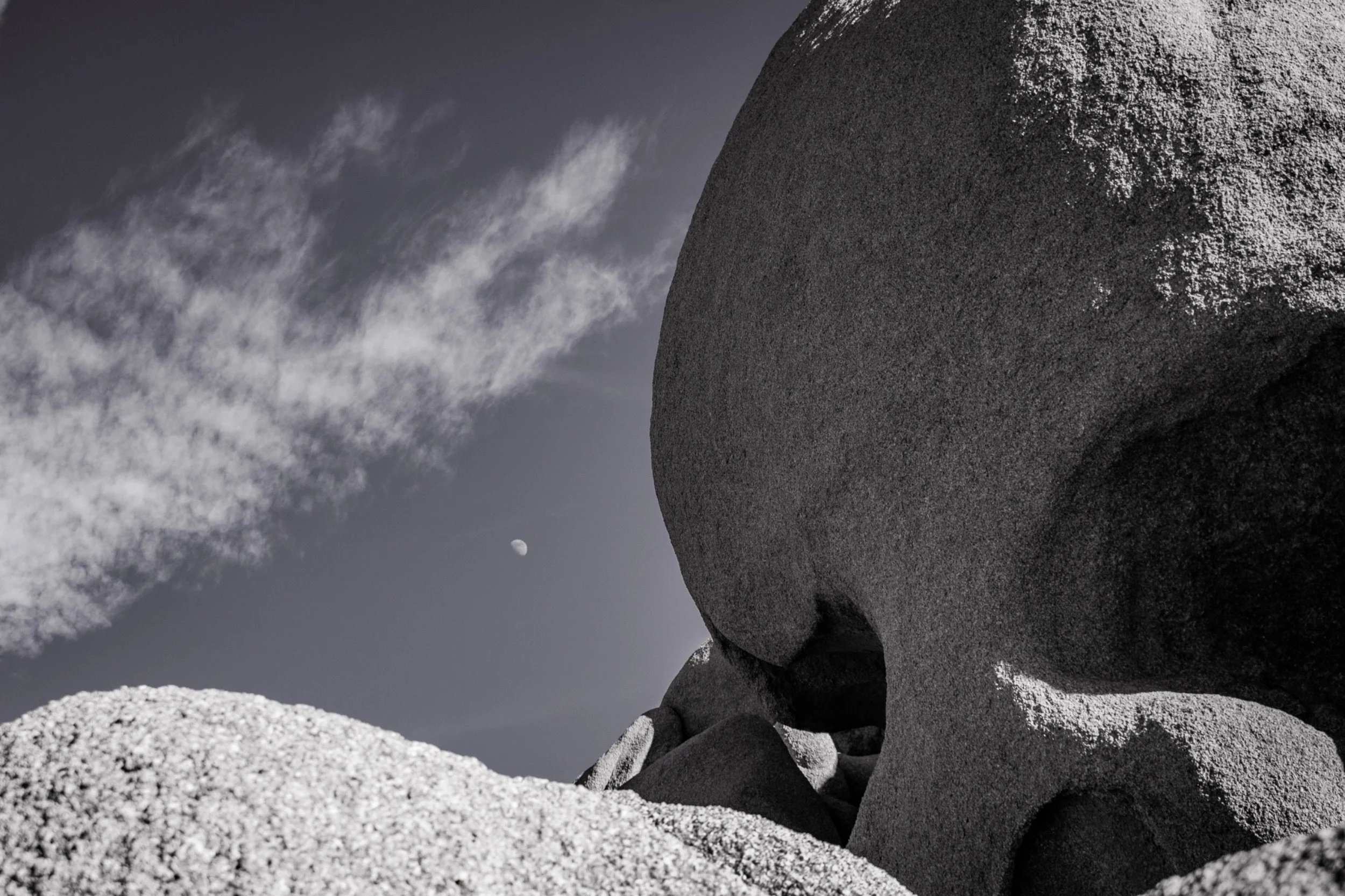Close-up view of rough, dark-colored rocks and boulders with a cloudy sky in the background. The moon is visible in the sky with wisps of clouds passing by.
