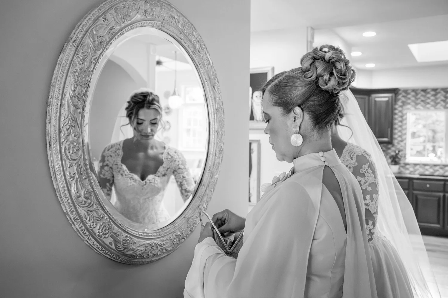 A bride with an updo hairstyle and lace wedding dress looks into a mirror while holding a phone, with a woman beside her. The reflection in the mirror shows the bride reading a message or looking down.