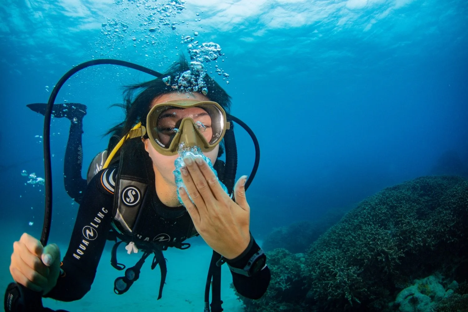 A woman scuba diving underwater, blowing bubbles, with coral and rocks in the background.