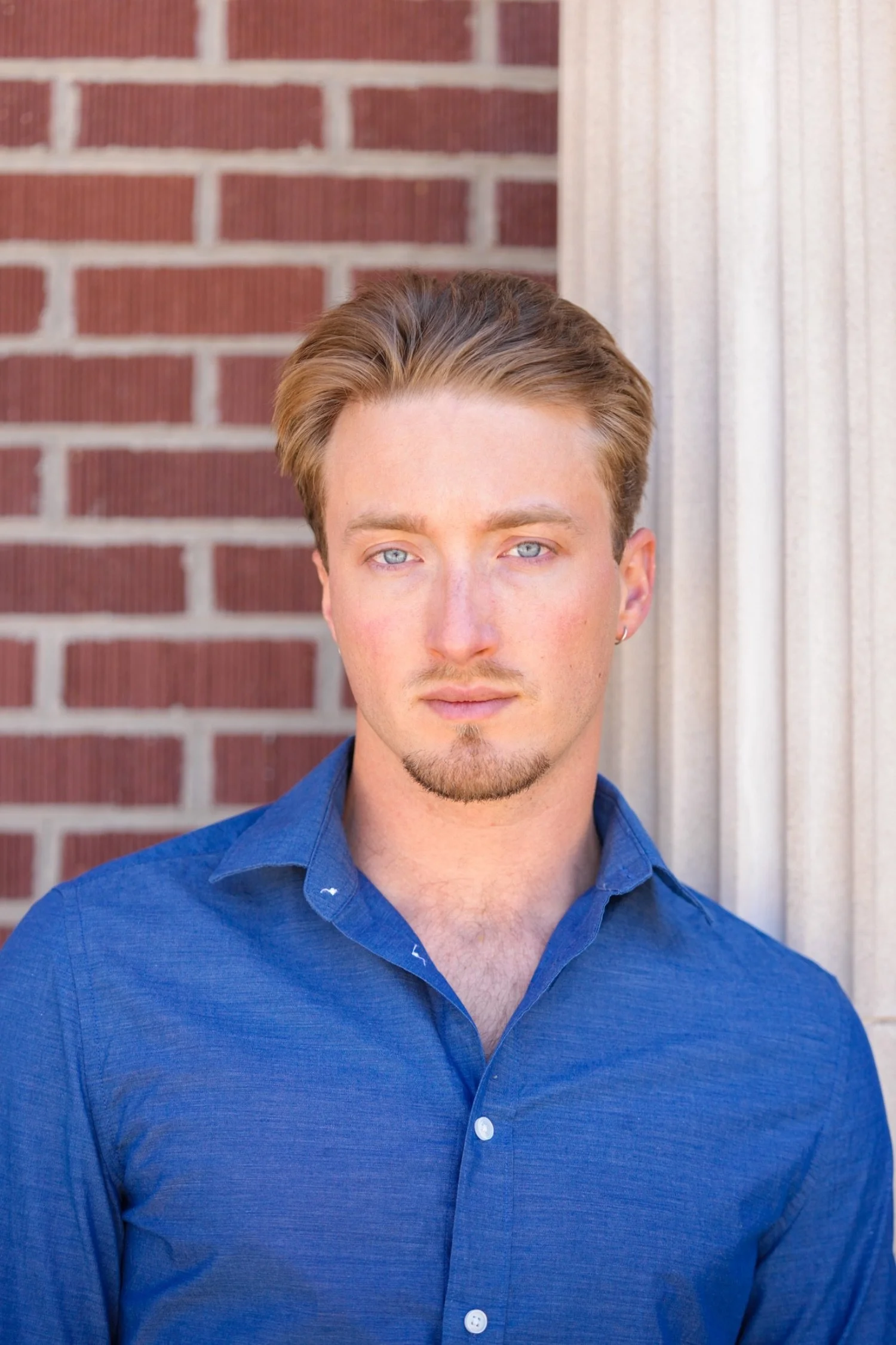 Close-up of a young man with auburn hair, blue eyes, and light skin, wearing a blue button-up shirt, standing in front of a brick wall and a white column.