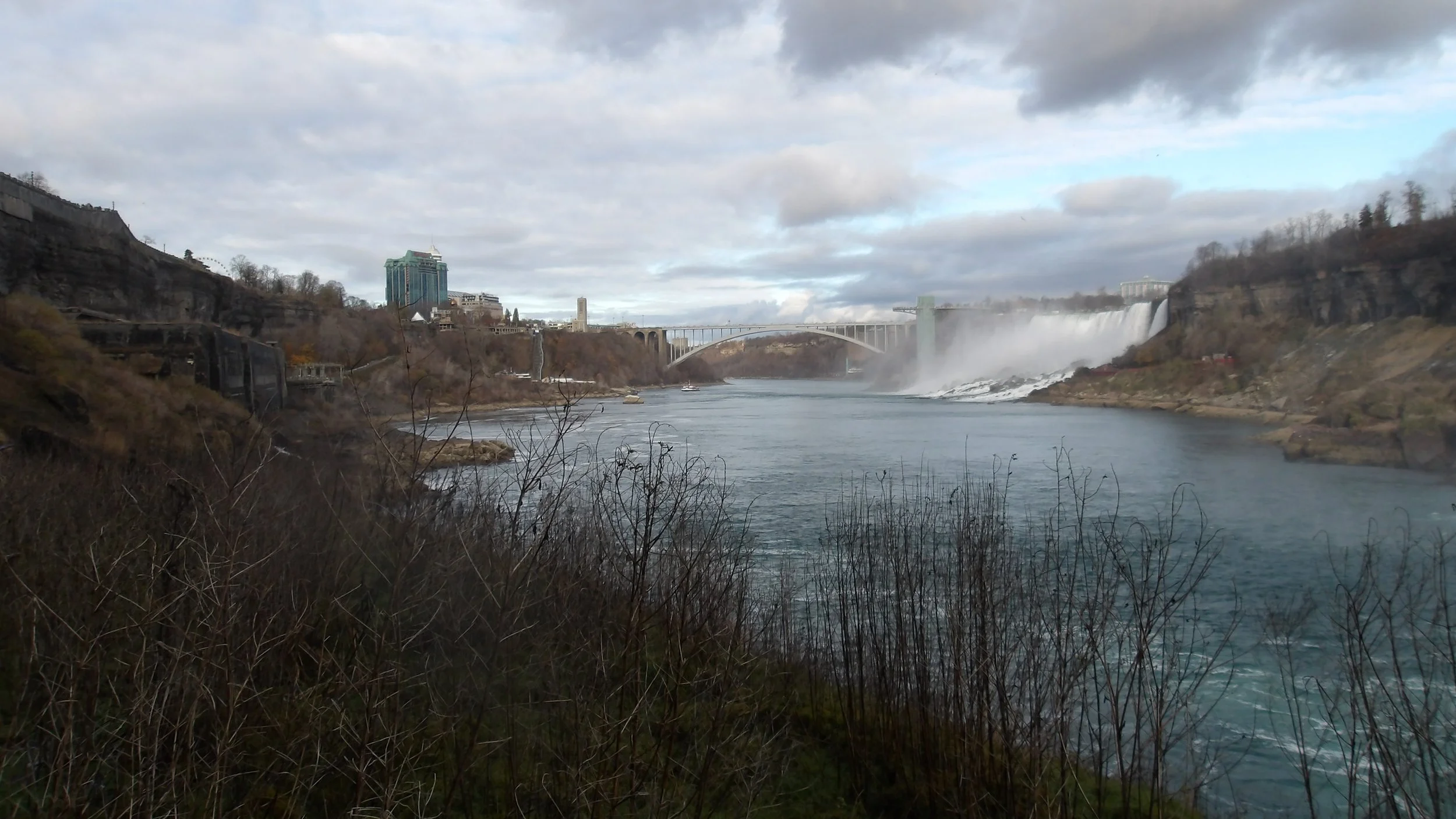View of Niagara Falls with the river in the foreground, mist rising from the falls, and city buildings in the background under a cloudy sky.