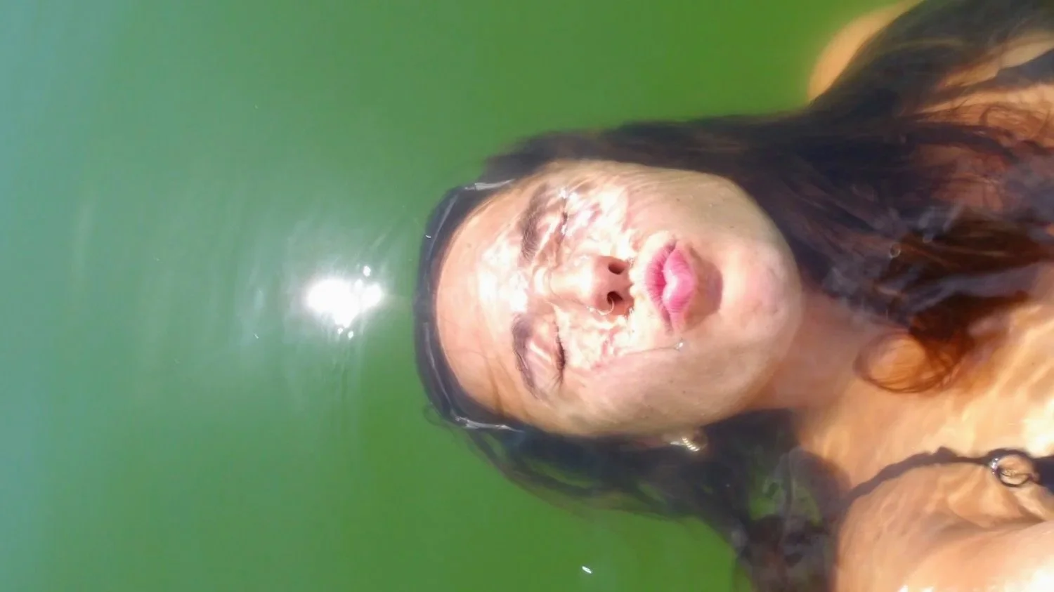 A woman with wet hair and glasses taking a selfie in a green body of water, her eyes closed and lips puckered.