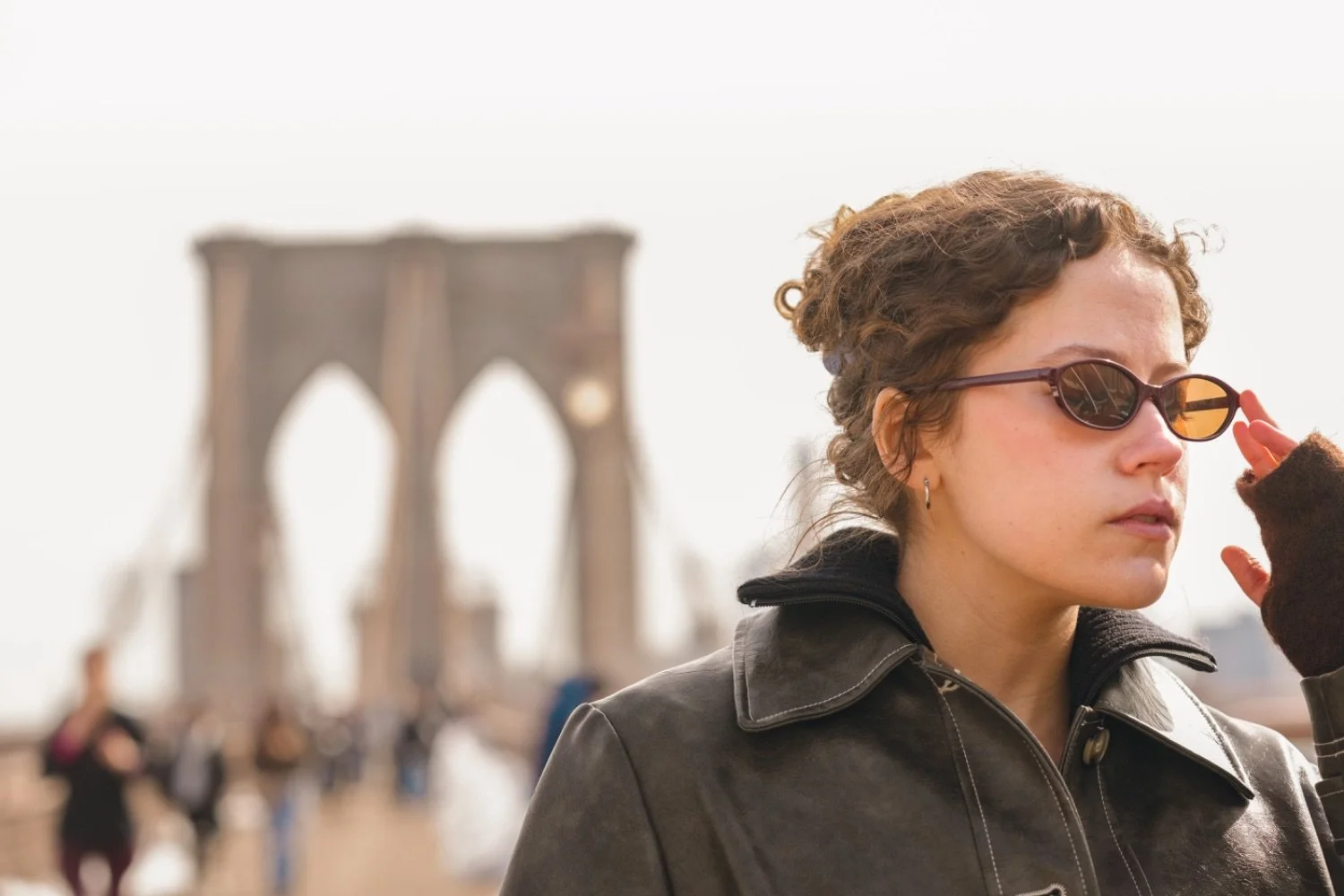 A woman with curly hair adjusting her sunglasses on a bridge with a blurred background of people walking, and a notable bridge structure in the distance.