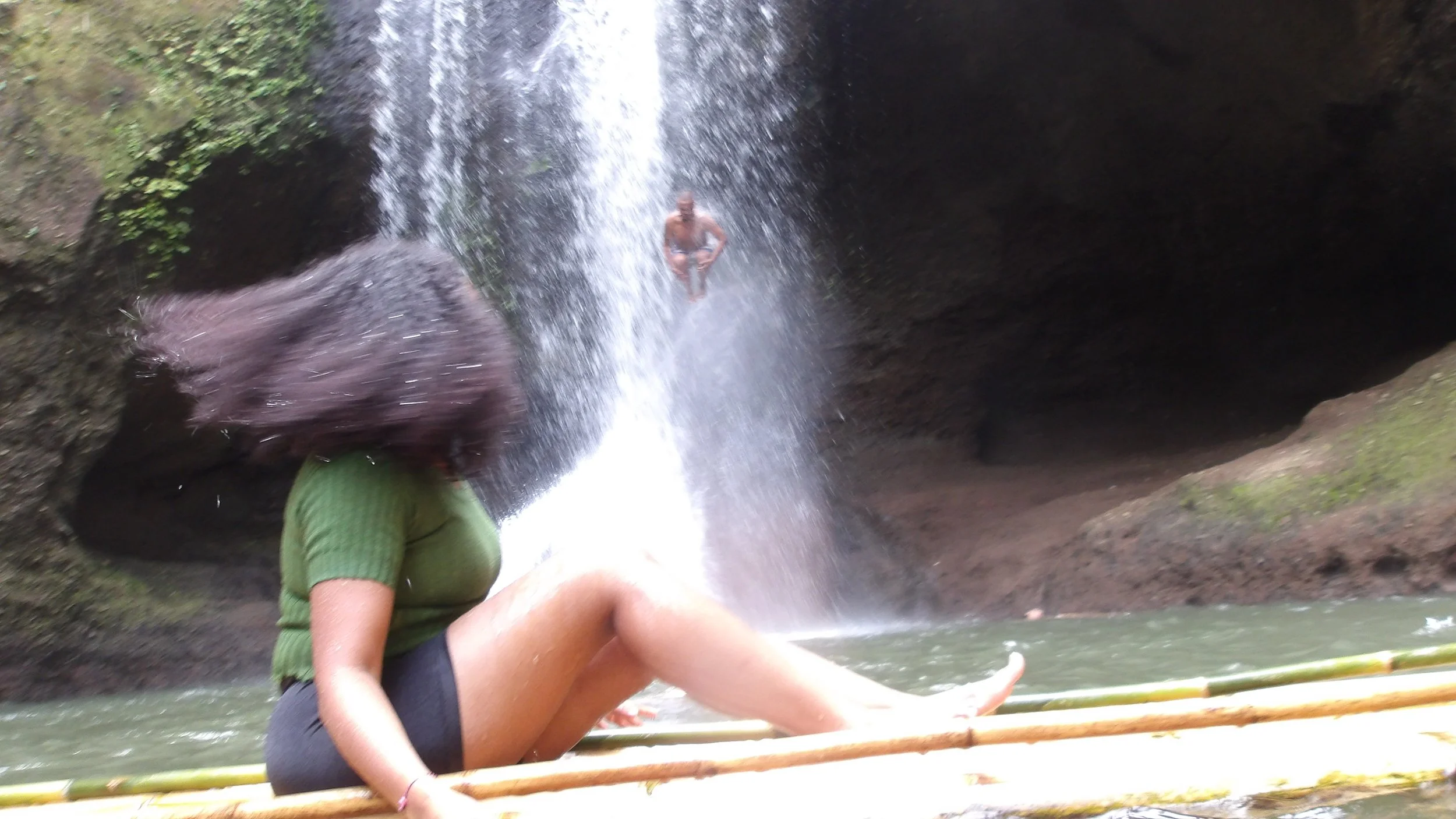 A person in a green shirt is sitting under a waterfall inside a cave, with another person visible in the background beyond the waterfall.