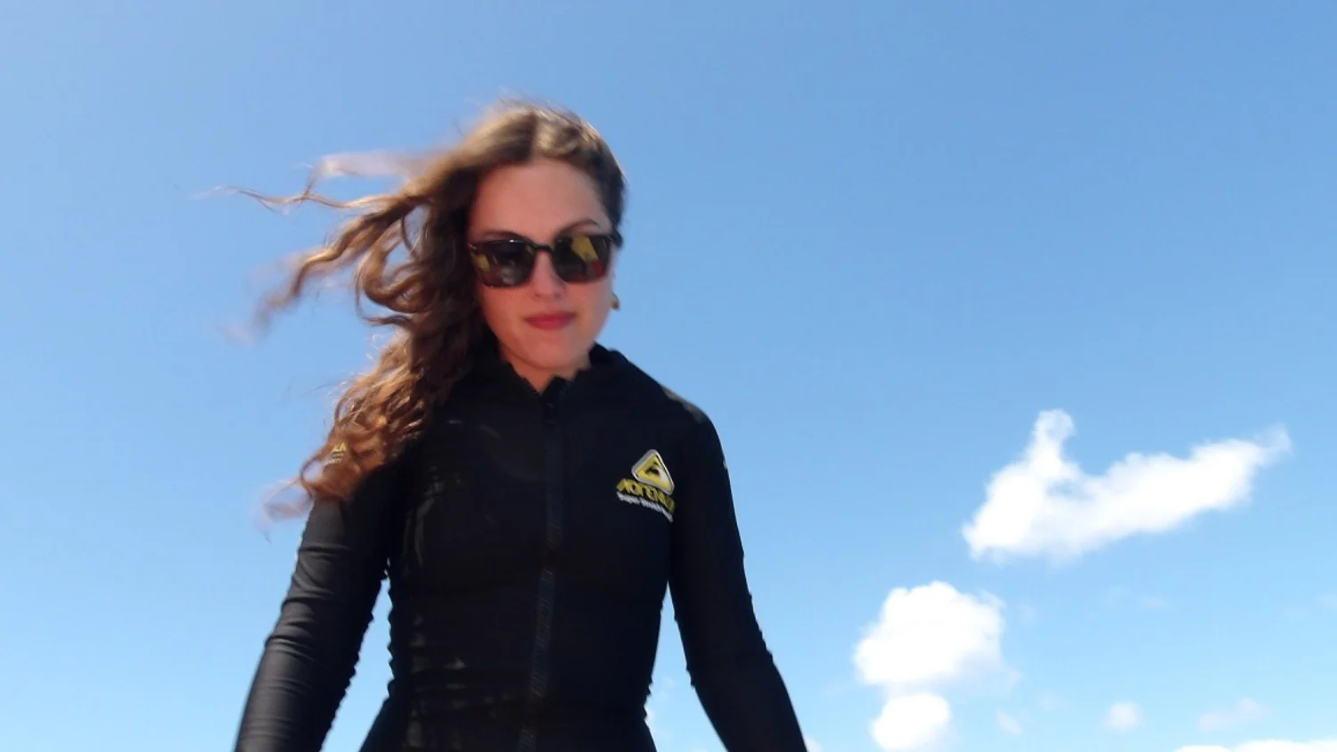 A woman with wavy brown hair, wearing sunglasses and a black jacket with a yellow logo, standing outdoors against a blue sky with a few clouds.