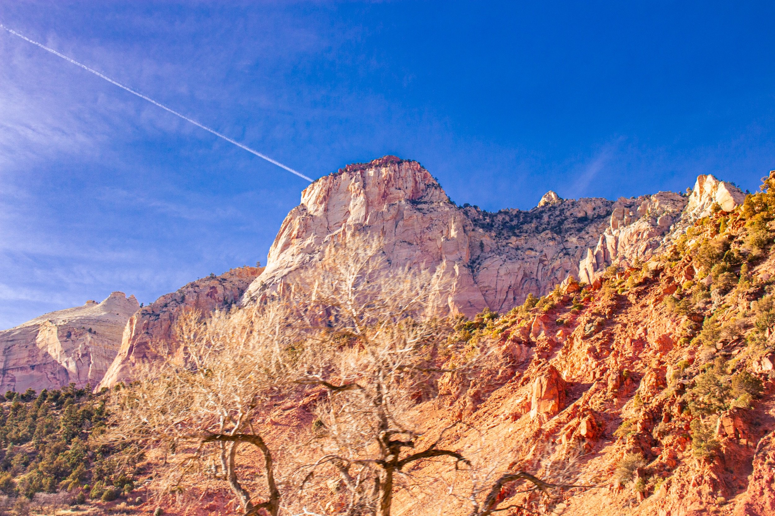 A mountainous landscape with large beige and orange-rock formations, sparse desert vegetation, and a vivid blue sky with a contrail.