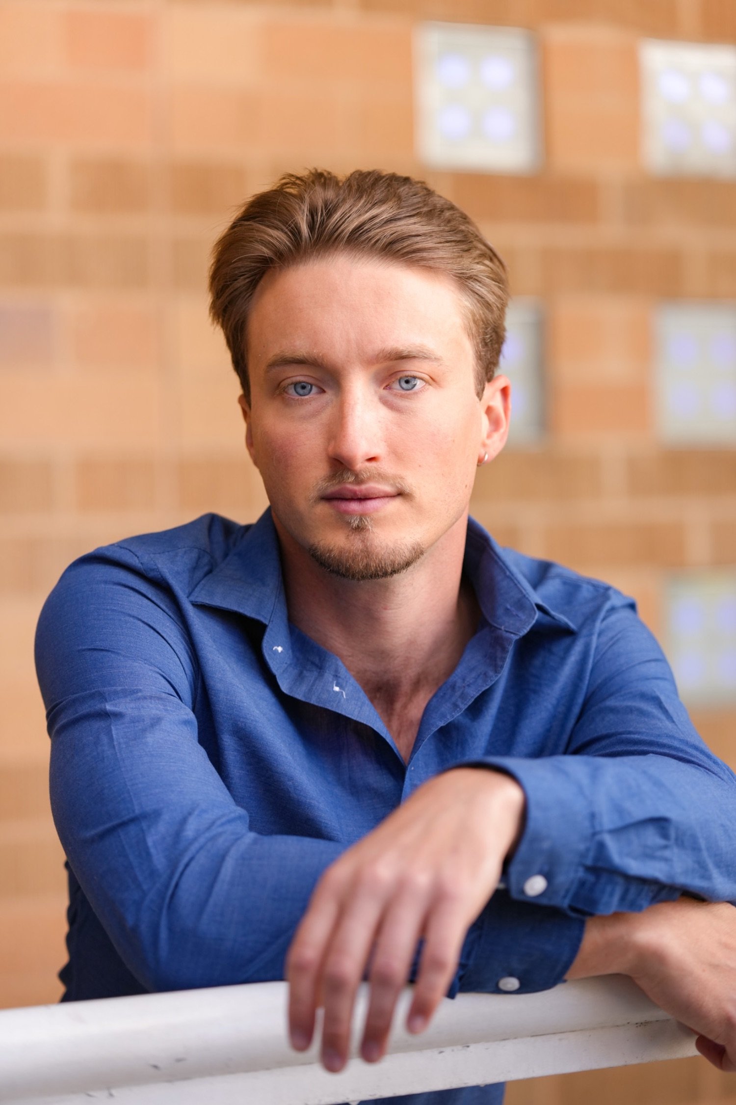 A young man with light skin, blue eyes, and brown hair, wearing a blue shirt, is sitting at a table with a brick wall and various framed pictures in the background. He is resting his right arm on the table, with his hand extended forward.