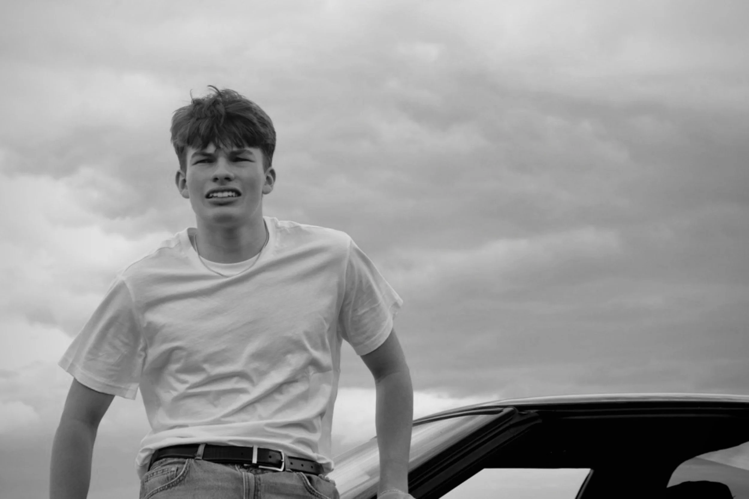 A young man with short hair wearing a white t-shirt and jeans, standing outdoors near a car, looking at the camera with a slightly concerned or puzzled expression against a cloudy sky.