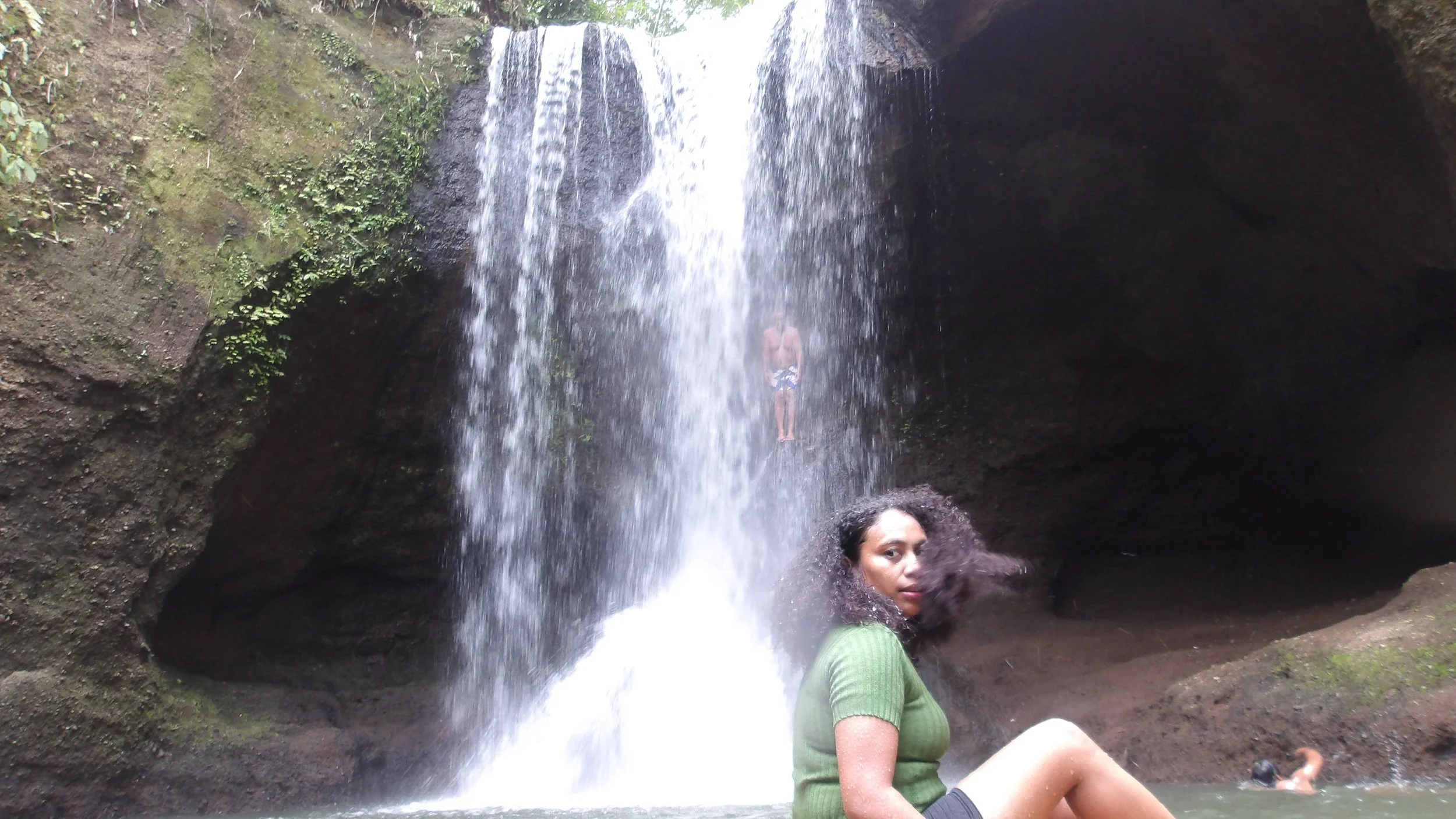 A woman with curly hair sitting in a small pool of water under a waterfall in a natural cave with moss-covered rocks and greenery.