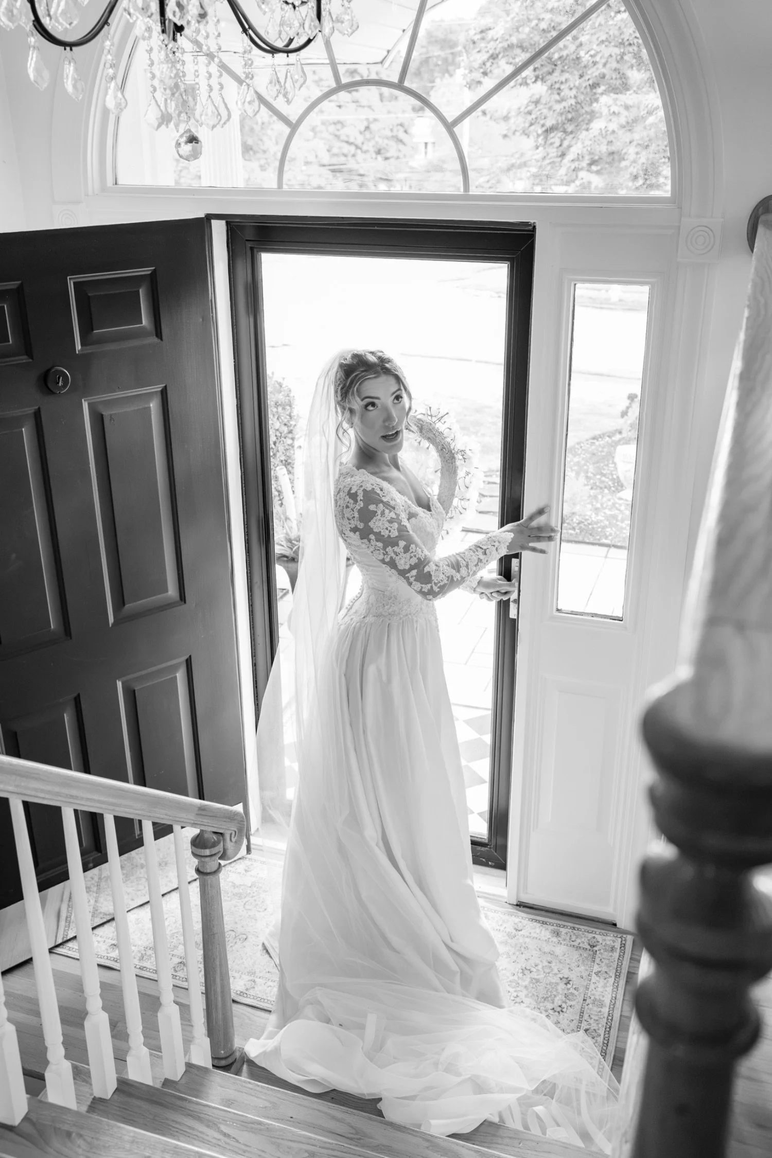 A bride in a lace wedding gown and veil standing in the doorway of a house, looking inside with a surprised or excited expression, as she prepares for her wedding.