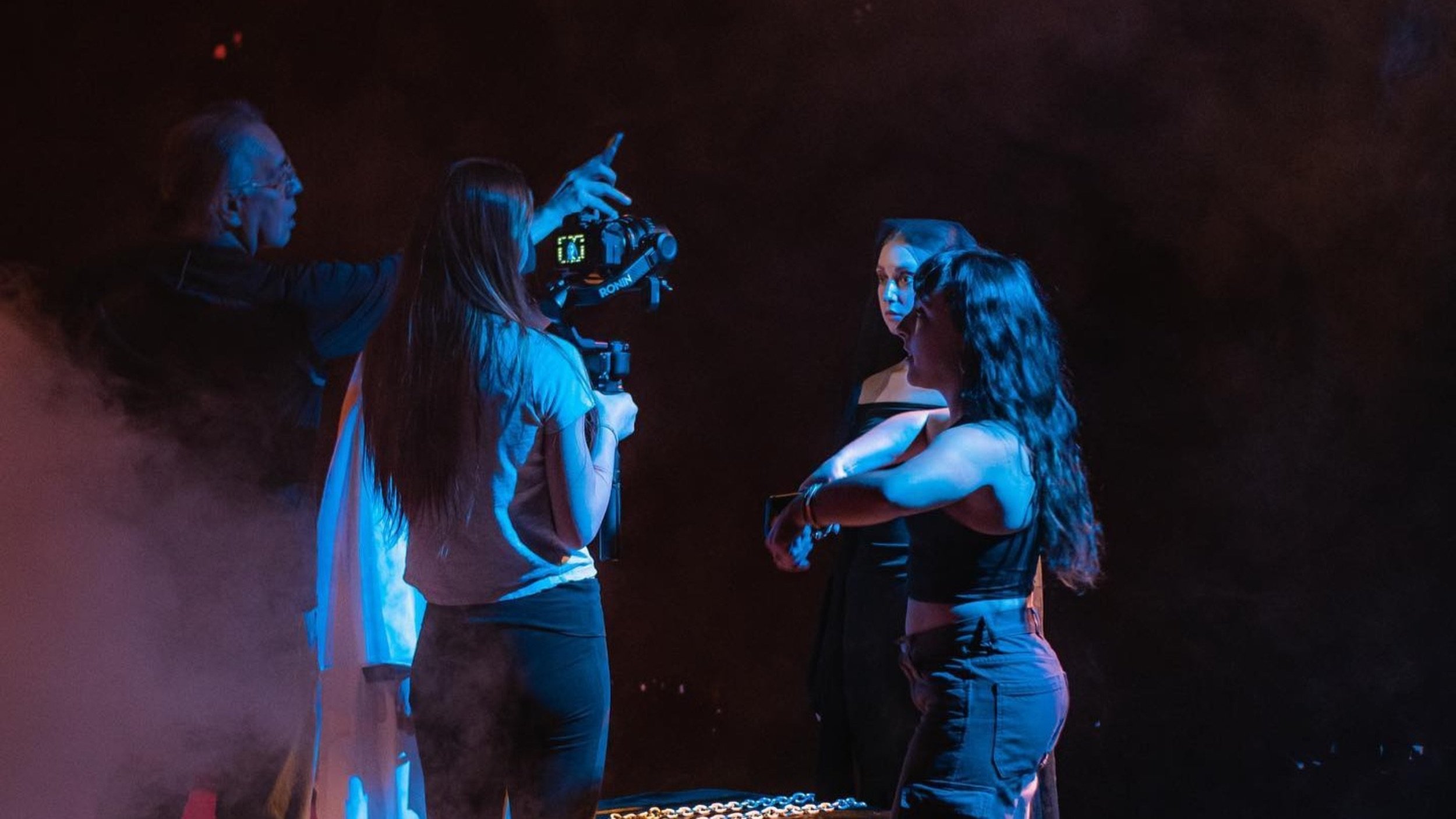 Four women are standing in a dark room illuminated by blue light, with one woman holding a camera on a tripod and the other three women engaged in conversation.